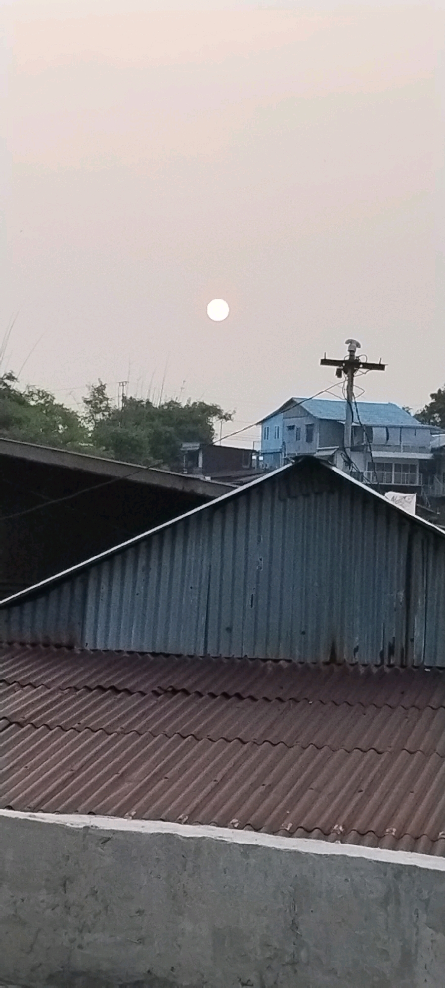 The image captures a hazy sky with a bright, white orb, possibly the sun or moon, positioned in the upper portion of the frame. In the mid-ground, several buildings are visible. One prominent building has a light blue roof and white walls, featuring multiple windows. To the left of this building, a utility pole with electrical wires is present. In the foreground, corrugated metal roofs of different colors, one appearing rusty brown and the other silvery-grey, dominate the lower half of the image, suggesting an urban or semi-urban setting. The overall lighting suggests it could be sunrise or sunset due to the diffuse quality of the light and the hazy atmosphere. The location, Balikpapan, Indonesia, implies a tropical or subtropical environment, which could contribute to the hazy conditions. There are no people or overt actions depicted in the scene; it is a static architectural and atmospheric view.