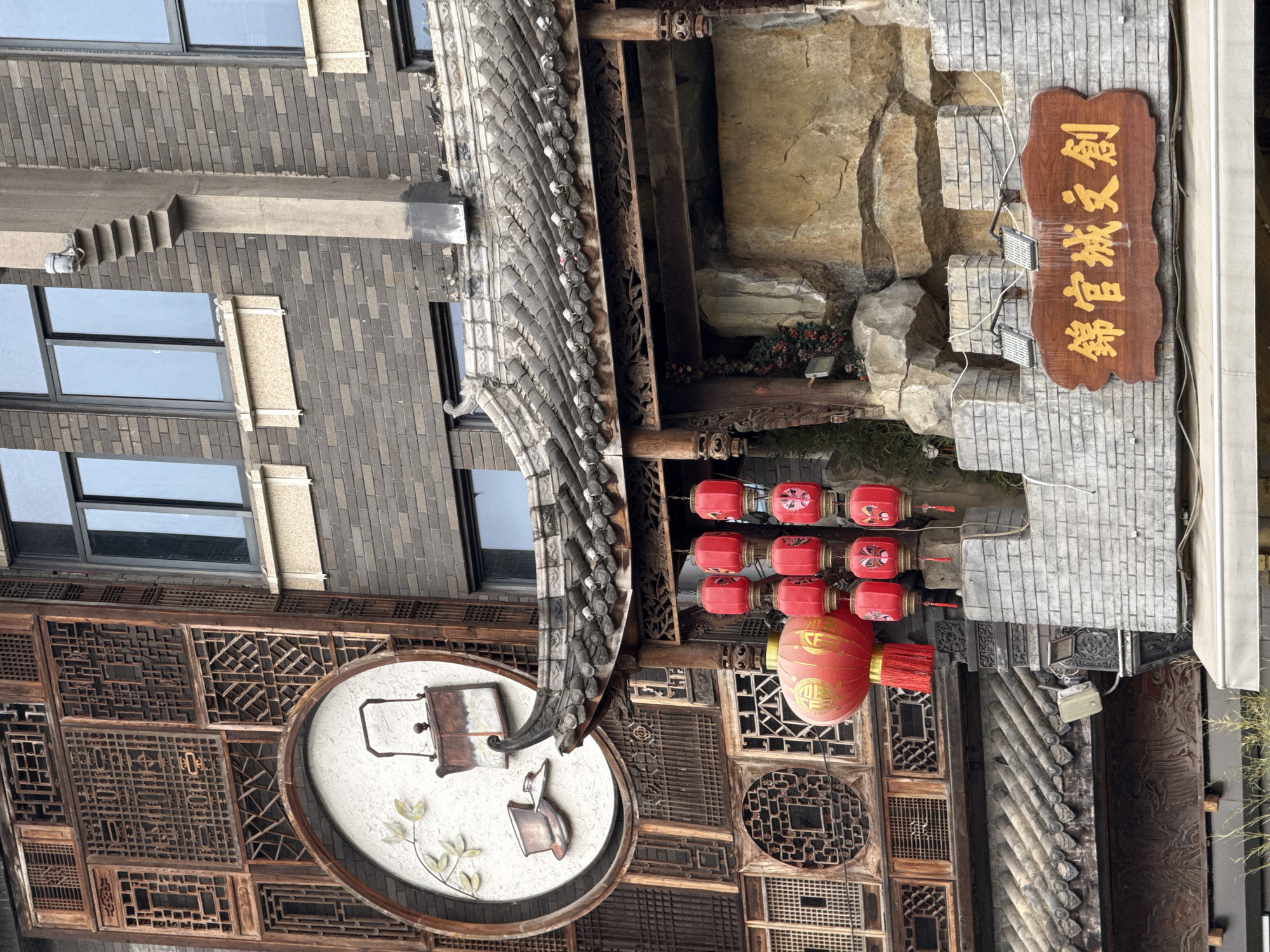 This image depicts a multi-story building facade in Qingyang District, China, showcasing a blend of traditional Chinese architectural and decorative elements with modern construction. The scene is devoid of people.
The left portion of the image features a traditional wooden facade composed of numerous intricate lattice panels with geometric and floral patterns, stained a reddish-brown. Centered on this facade, at mid-height, is a large circular white emblem with a raised relief depicting a traditional Chinese teapot, a teacup, and a branch with light green leaves.
To the right, a modern building constructed with dark grey bricks is visible, featuring rectangular windows with black frames and light-colored window sills. A vertical, light grey concrete column with a stepped, inverted-pyramid design extends upwards from just above a traditional roofline, reaching toward the upper windows of the brick building. A small metallic fixture, possibly a light or camera, is mounted on top of this concrete column.
Below the concrete column and the wooden facade, a traditional Chinese-style roof with dark grey, ridged tiles and ornate eaves juts out. The eaves are supported by intricately carved wooden pillars and beams. From beneath this roof, a cluster of red Chinese lanterns hangs. This cluster includes one larger spherical lantern with gold Chinese characters and a red tassel, and several smaller, cylindrical lanterns decorated with painted facial masks, arranged in vertical stacks.
Beneath the traditional roof, a facade designed to mimic natural rock formations is present, with varying shades of grey and beige, interspersed with small artificial greenery. At the bottom of the image, a grey brick wall, designed with crenellated battlements at its top, supports a rectangular wooden sign. The sign features raised gold Chinese characters that read "錦官城文創" (Jǐn Guān Chéng Wén Chuàng). Two small light fixtures are mounted on the grey brick wall, positioned to illuminate the wooden sign.