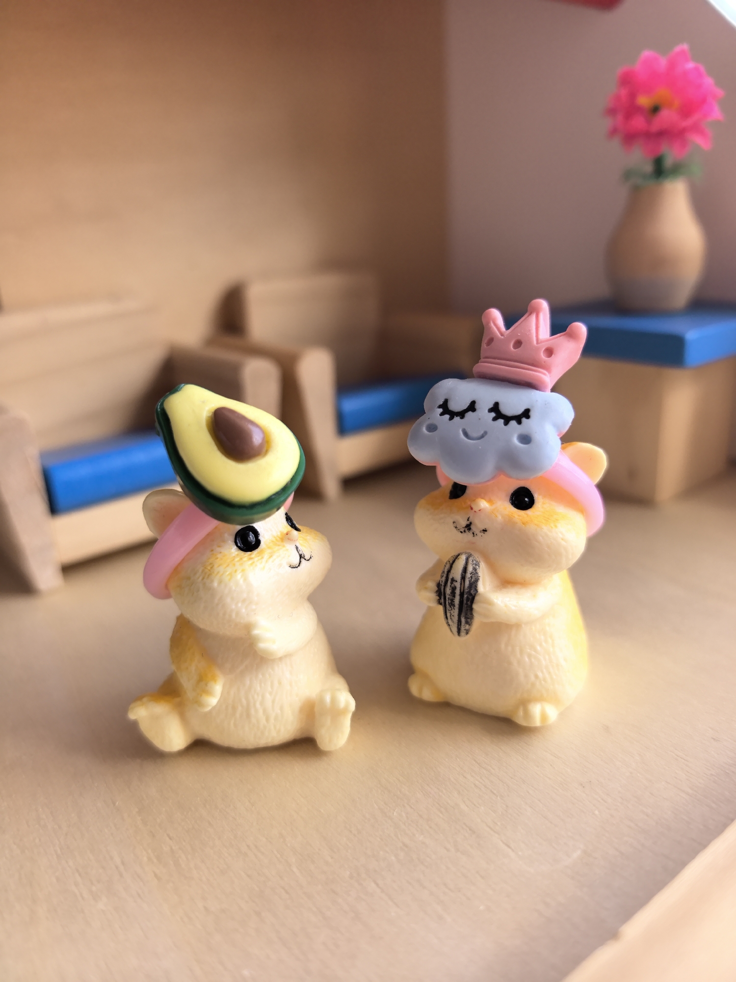Two miniature hamster figurines are positioned within what appears to be a dollhouse setting. The left figurine features a slice of avocado balanced atop its head, secured by a pink headband. The right figurine has a blue cloud with closed eyes and a pink crown on top, also secured by a pink headband. The right figure is holding a sunflower seed. The background includes miniature furniture: a couch with blue cushions and a shelf with a vase containing a pink flower. The location is not discernible from the image.