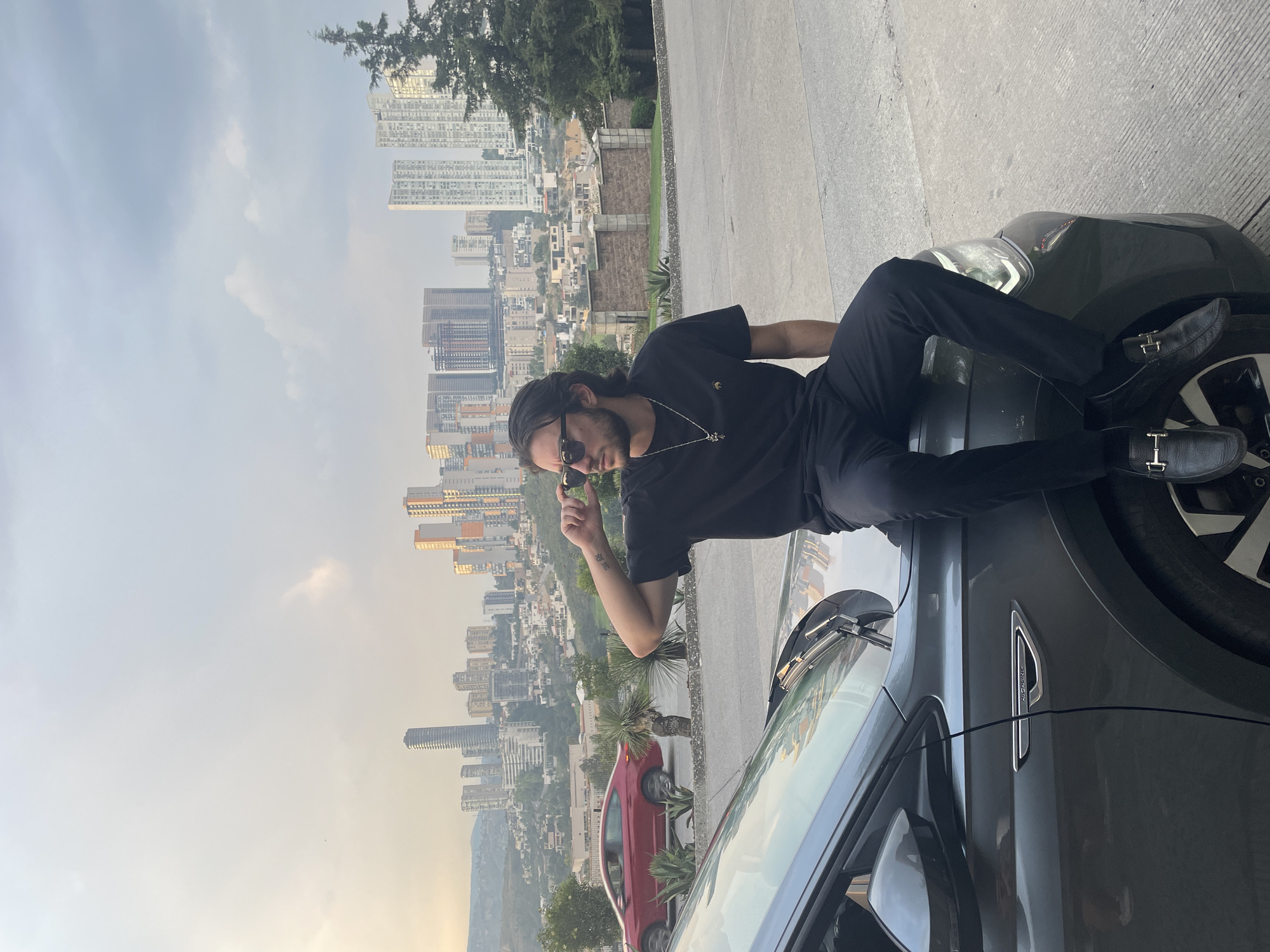 A man in dark clothing is reclining on the hood of a gray car, resting his legs on the front windshield and tire of a darker green vehicle. He is wearing sunglasses and a silver necklace. In the background, a cityscape is visible under a cloudy sky. A red car is parked near the vehicles. The location is Huixquilucan de Degollado, Mexico, suggested by the urban skyline.