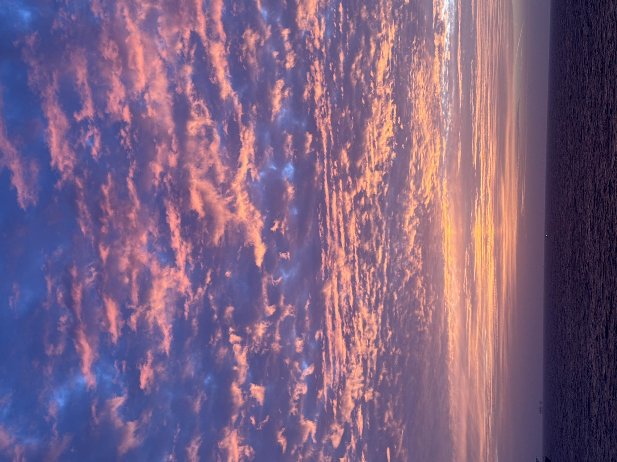 The image shows a sunset over the ocean. The sky is filled with textured clouds that range in color from blue to pink to orange. The sun is obscured by the clouds, but its rays are visible. The ocean is dark and calm. The location is unknown.