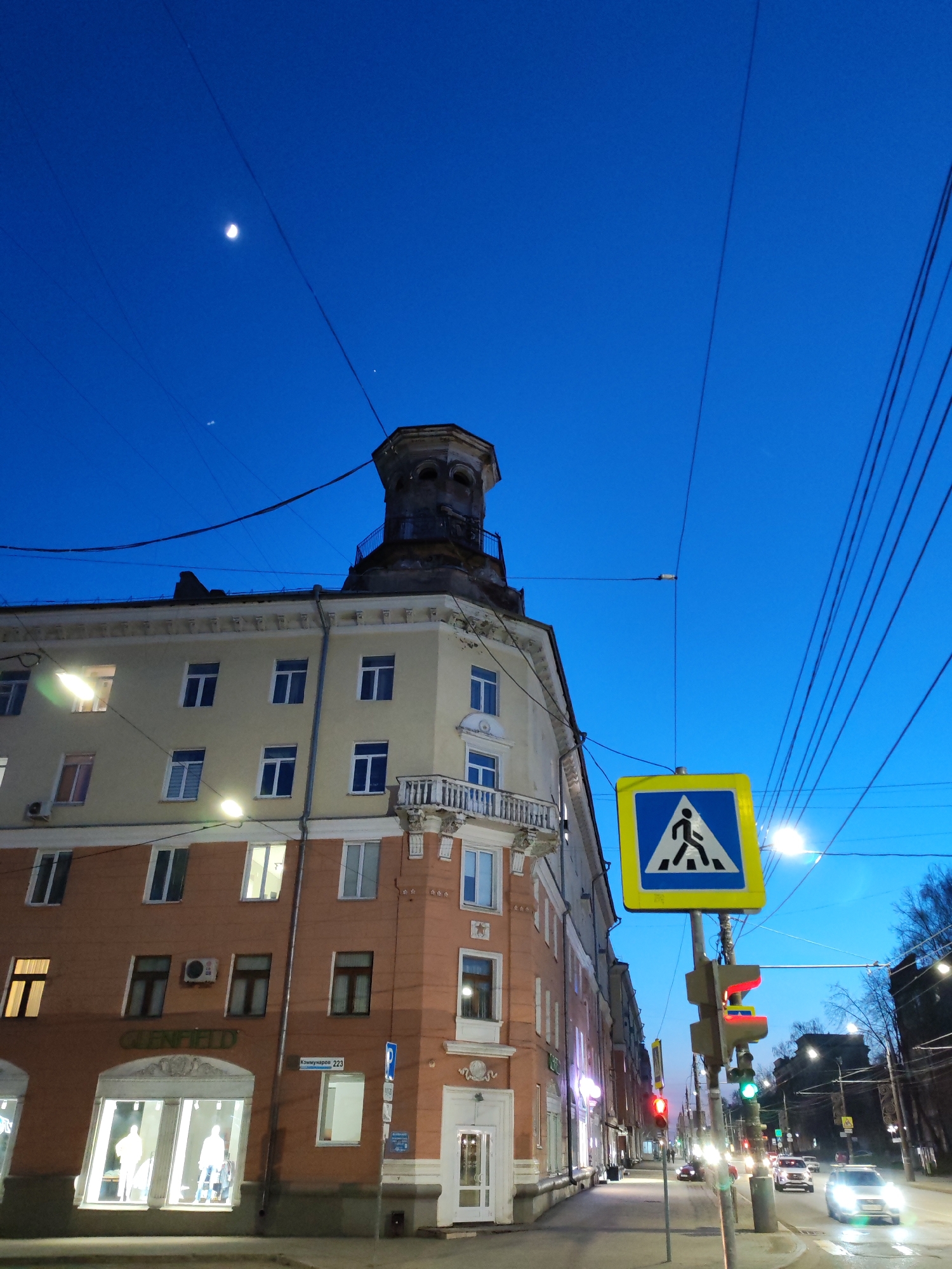 This media file presents an urban street scene in Izhevsk, Russia, captured during dusk or early night. The sky is a deep, clear blue, featuring a distinct crescent moon in the upper left quadrant and a fainter light source nearby. Numerous overhead power lines traverse the sky, adding to the urban feel.
The primary subject is a multi-story building situated on a street corner, its upper levels a light yellow with several windows, contrasting with a reddish-brown lower facade. A distinctive, somewhat weathered, ornate tower or cupola caps the building's corner. The ground floor houses storefronts, one of which is a "GLENFIELD" clothing store, identifiable by its green logo and brightly lit display windows showcasing mannequins. A street sign affixed to the building clearly reads "Коммунаров 223" (Kommunarov 223), indicating its address, alongside a blue parking sign with a 'P' symbol.
In the foreground, a prominent blue pedestrian crossing sign featuring a white walking figure stands beside a set of traffic lights. The vehicular traffic light displays red, while the pedestrian signal shows green. A paved road stretches into the distance, with several cars visible, their headlights illuminated, suggesting active evening traffic. Streetlights provide bright artificial illumination, highlighting parts of the sidewalk and buildings. The scene captures the transition from day to night in a bustling city environment.