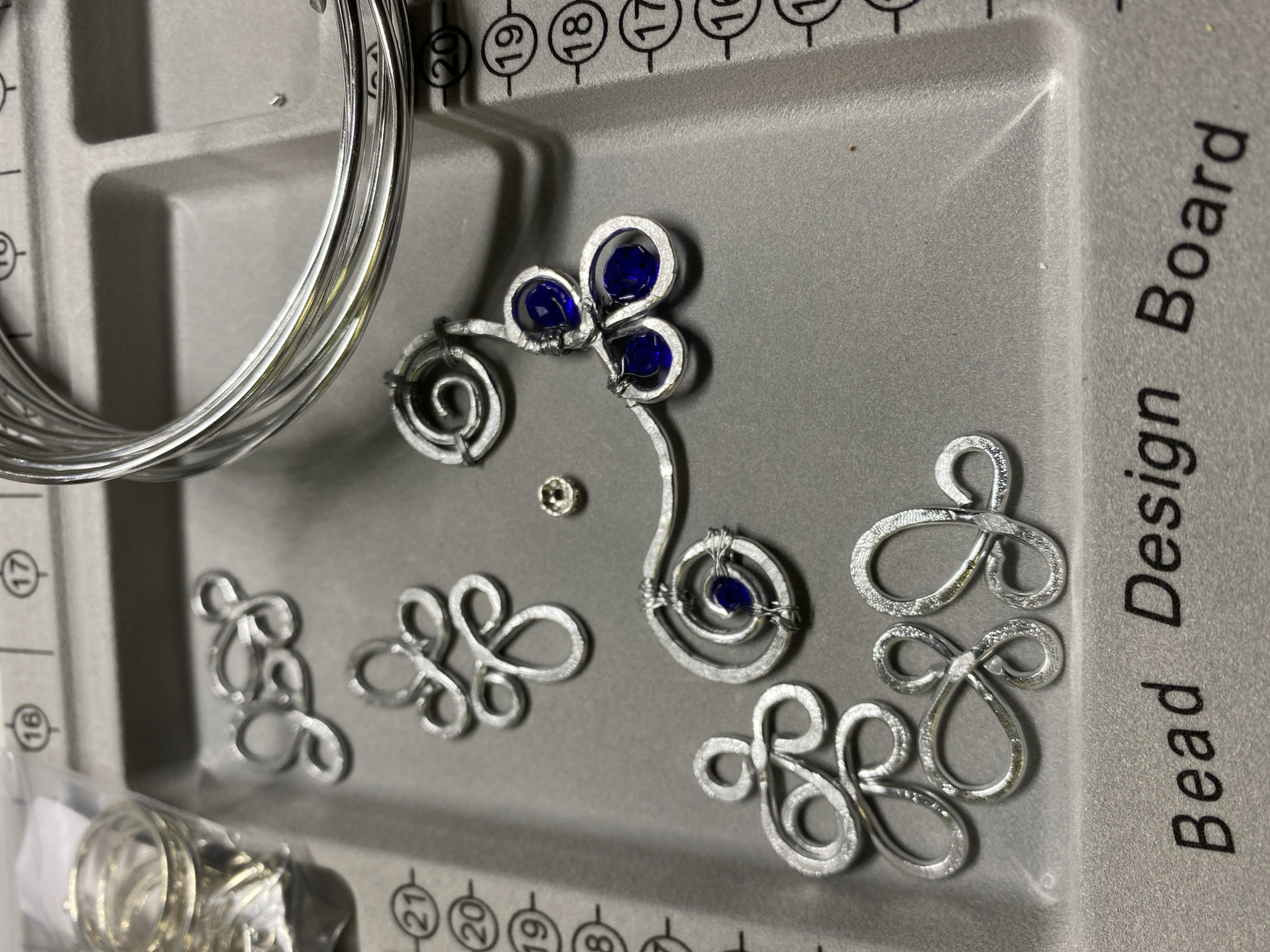 The image shows a close-up overhead view of a grey bead design board with various jewelry-making components. In the upper left corner, a plastic bag contains a stack of silver-colored rings. To the right of the bag are several silver-colored wire ornaments, one with three blue gemstone accents. A spool of thin silver wire is prominently displayed in the upper right quadrant of the image. The board itself has markings with numbers, indicating measurements, with numbers 16 through 21 visible. The text "Bead Design Board" is printed along the bottom edge of the board. There are no people or animals visible in the image, and no indication of a specific city or location, including Kushchovskaya, Russia.