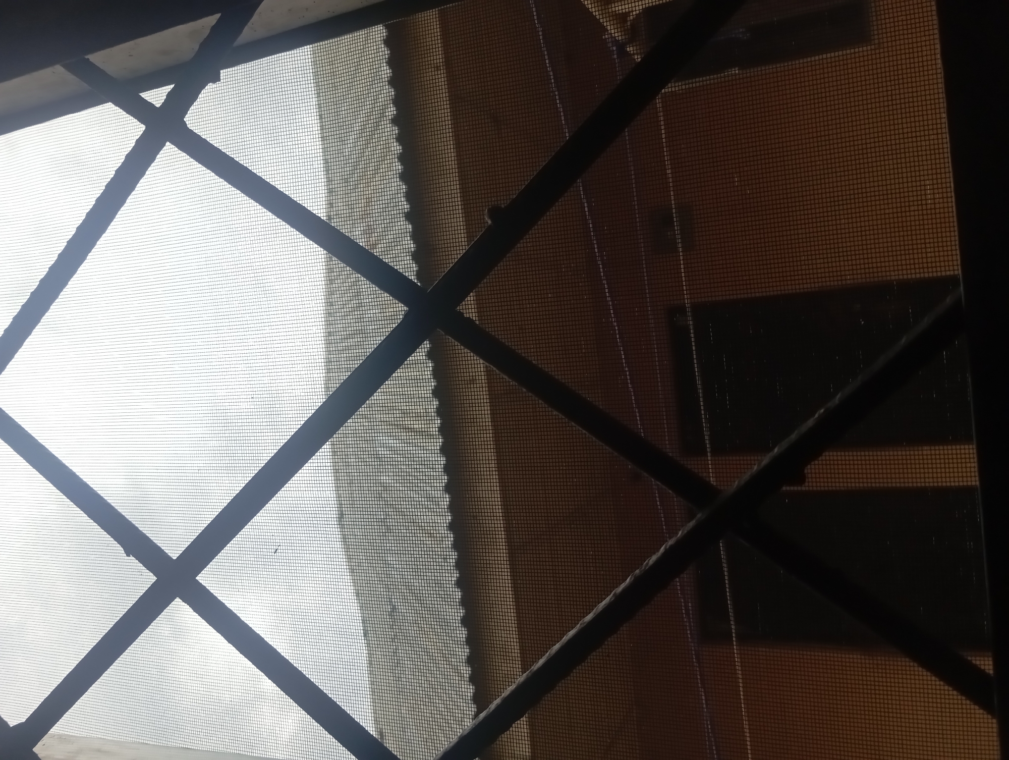 The image depicts a view through a window with a dark metal criss-cross security grate and insect screen. Beyond the grate, a building with a corrugated roof is visible. The building's walls appear to be painted a muted orange or tan color. Two dark, rectangular shapes, possibly windows or doors, are discernable on the building's facade. The sky above is overcast and bright, suggesting it is daytime. The scene is framed by the darker interior of the room from which the photograph is taken, particularly on the right and bottom edges. The overall impression is one of a somewhat utilitarian or aged urban environment.
