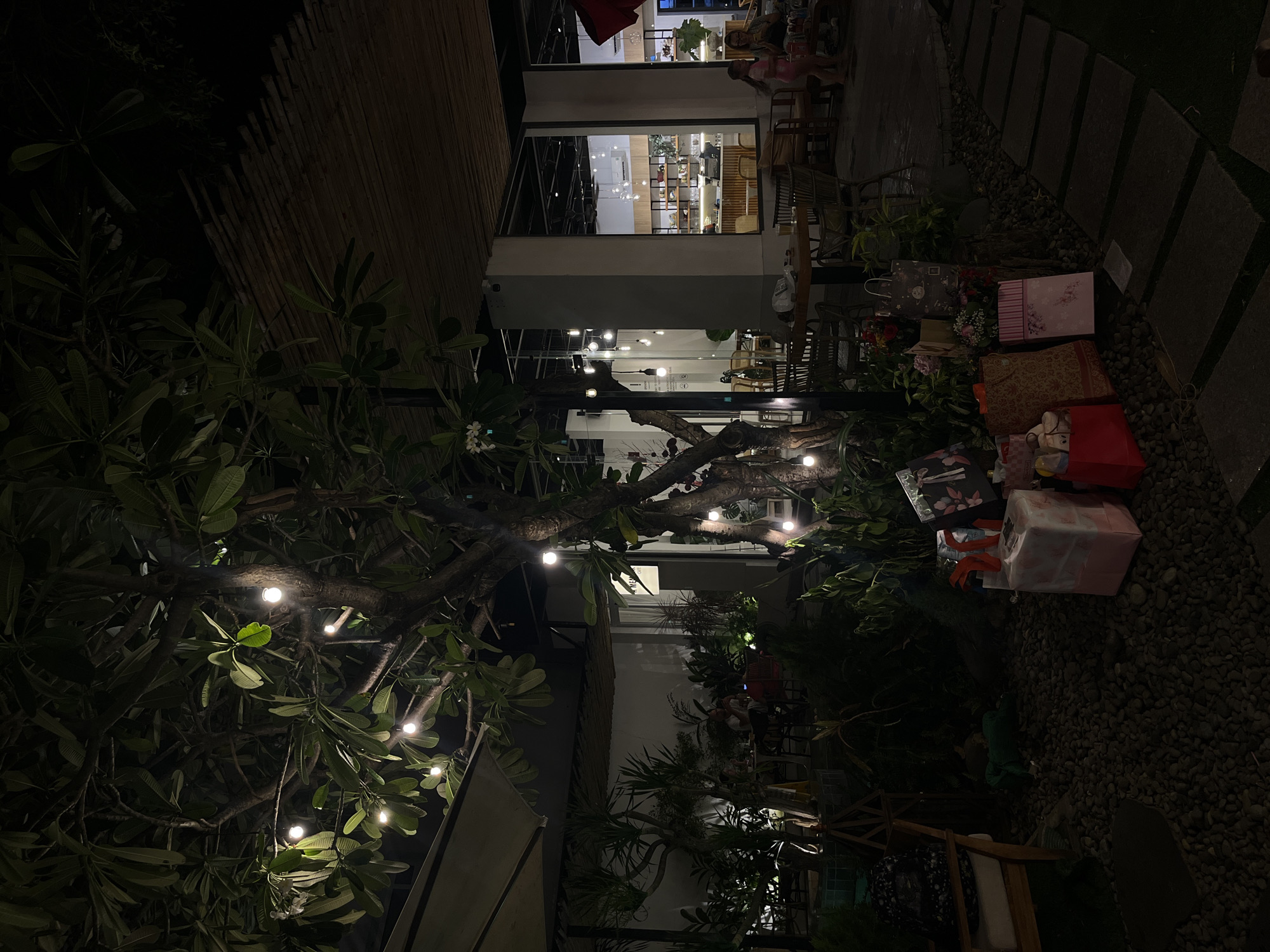 The image depicts an outdoor seating area of a cafe at night, with a tree adorned with fairy lights in the foreground. Several gift-wrapped packages are arranged on the ground near the tree. In the background, the cafe interior is visible through large glass windows, with patrons seated at tables. A woman and a child are standing near a table on the right side of the image. The scene appears to be located in Nam Nha Trang Ward, Vietnam.