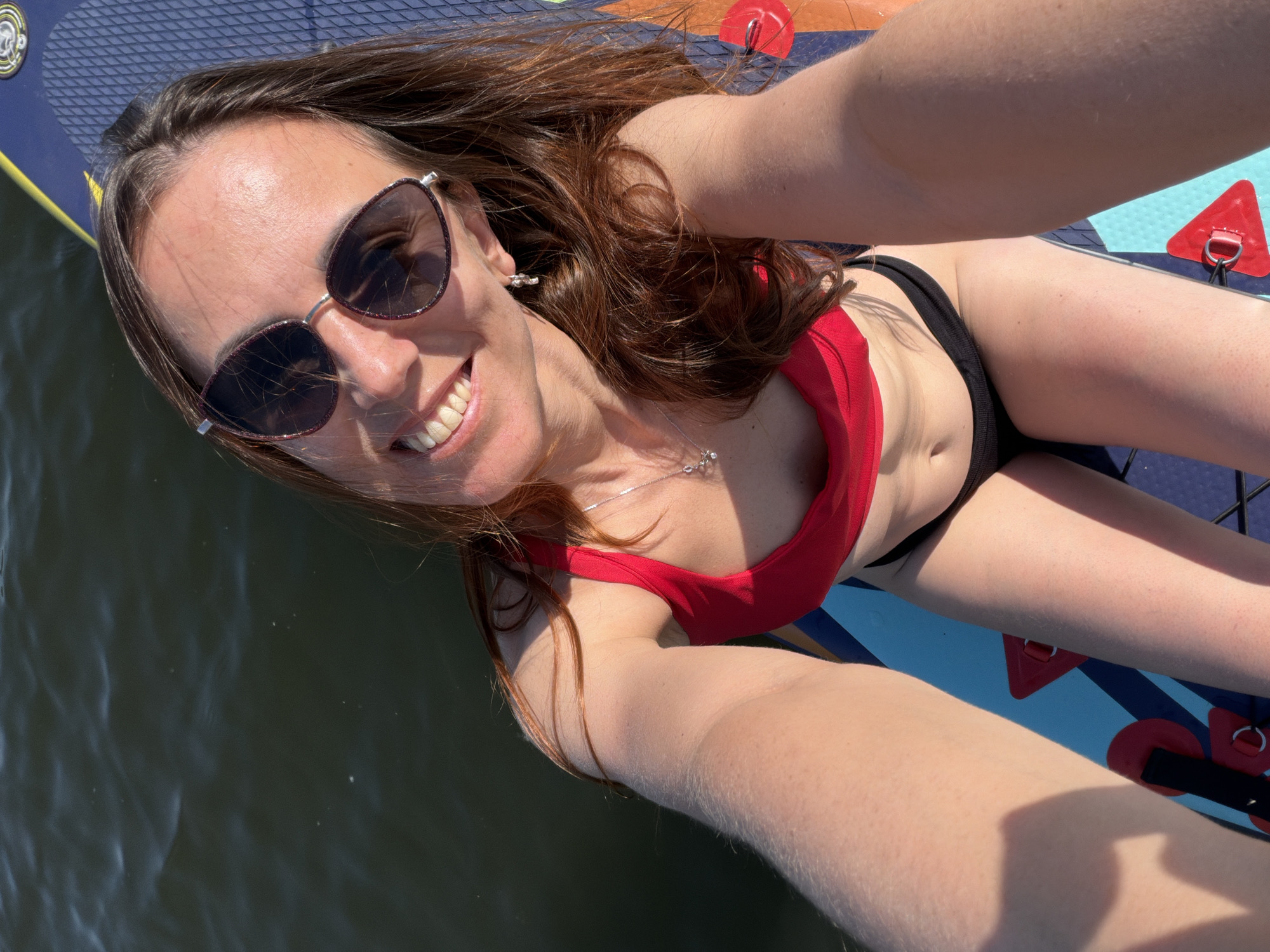 A woman is taking a selfie on a paddleboard in a body of water. She is wearing sunglasses and a red top with black bottoms. The paddleboard is blue and white with red accents. The water is dark and rippled. There is no indication of the location being Birobidzhan, Russia.
