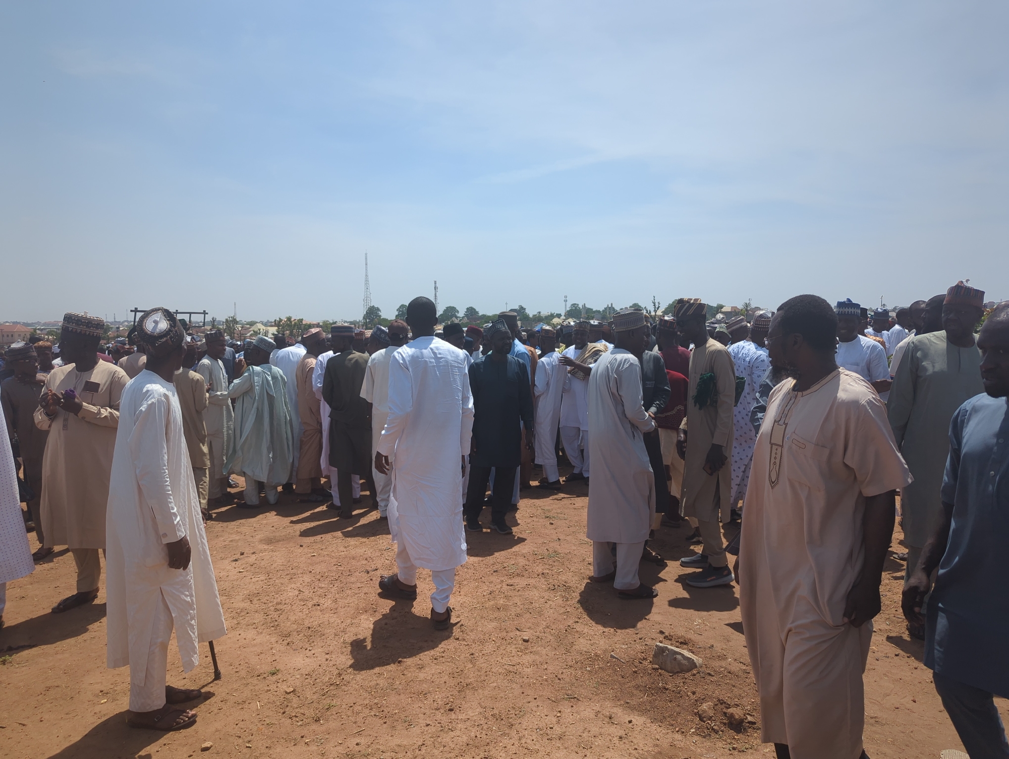 This image depicts a large outdoor gathering of men under a clear, bright blue sky, likely in Sardauna, Nigeria. The scene is dominated by a dense crowd of adult males, many of whom are dressed in traditional West African attire, including long, flowing robes (kaftans or boubous) and embroidered skullcaps. The clothing varies in color, with shades of white, beige, light blue, and darker hues visible.
The ground is unpaved, characterized by reddish-brown earth, suggesting a dry or semi-arid environment. Strong sunlight from directly overhead casts distinct, short shadows from the individuals onto the ground, indicating it is likely midday or early afternoon. In the foreground, one man in a white robe and sandals is partially visible from behind, leaning on a black cane.
The men are standing closely together, forming a large congregation, with their bodies generally facing inward or around. Their expressions are not clearly visible due to the distance and angles, but the atmosphere appears calm. In the background, beyond the crowd, several buildings and multiple communication towers can be seen, suggesting the gathering is taking place within or on the outskirts of a town or city. No visible text is present in the image.