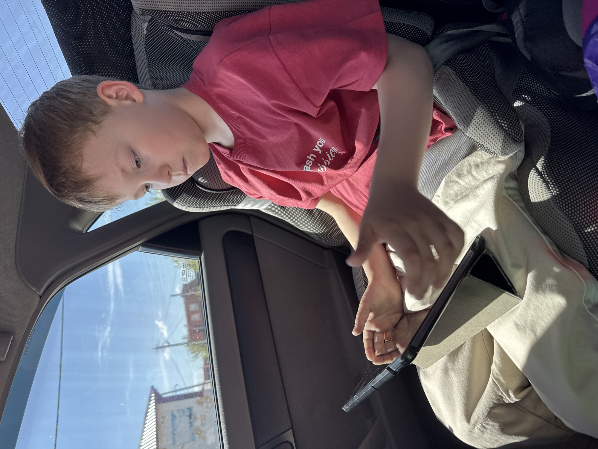 A young boy with fair hair is seated in a car, presumably in the back seat, as evidenced by the angle and the presence of a window to his left showing the outside. He is wearing a red t-shirt and khaki pants, and he is looking down at a tablet computer that he is holding with both hands. The surrounding environment is the interior of a car, with the car door and seat visible. The light suggests it is daytime, and through the car window, buildings and power lines are visible. It is not possible to confirm if this is Balezino, Russia from the image.