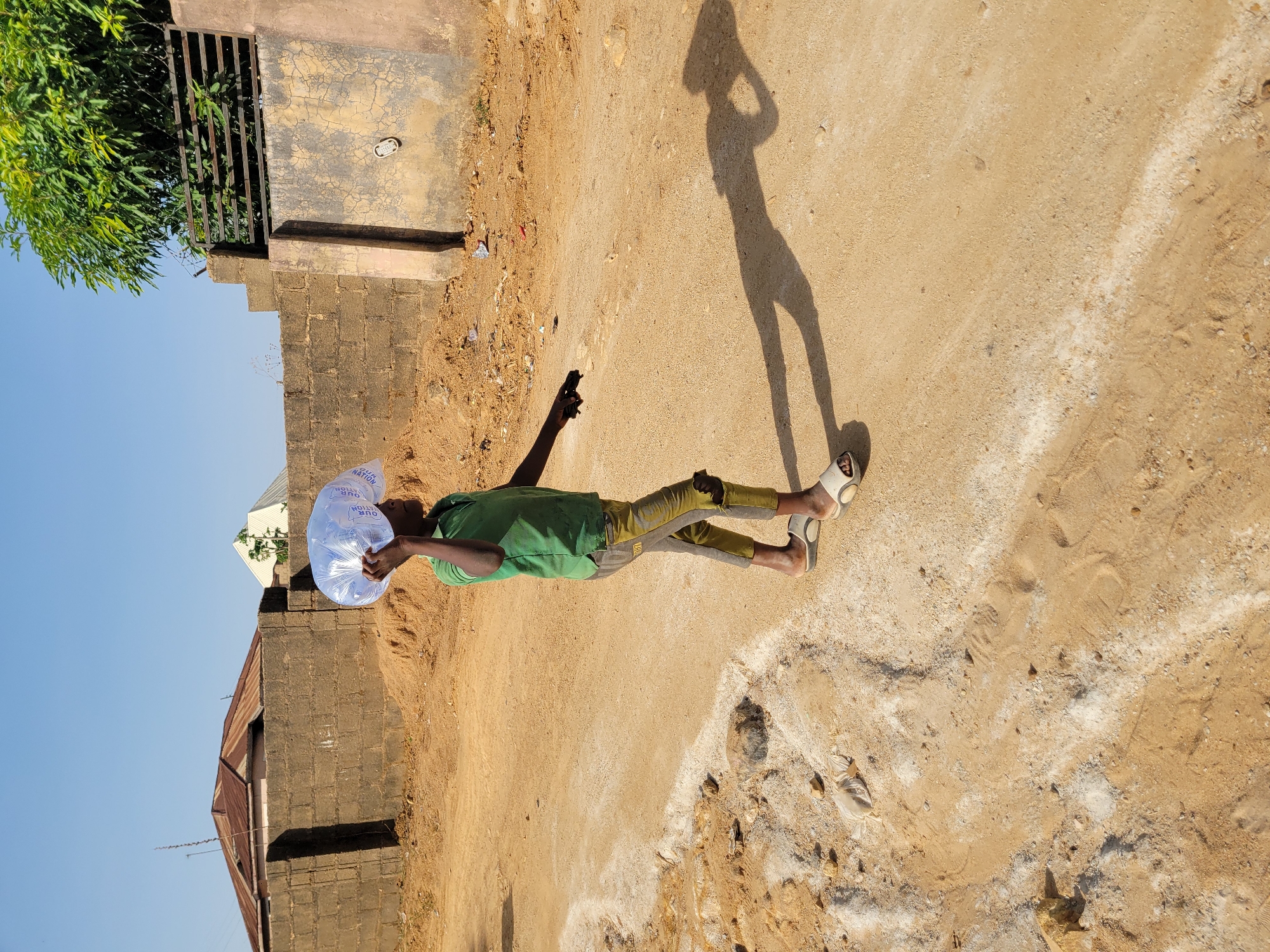 This image, captured in Barakin Akawo, Nigeria, depicts an outdoor scene on a sunny day. The central subject is a person, likely male, dressed in a green t-shirt, greyish-yellow trousers, and white and black sandals. They are standing or walking on a light brown dirt path.
The person holds a translucent white plastic bag, possibly filled with water or other items, over their head with their left hand, perhaps for shade or to carry it. In their right hand, held down by their side, they hold a black smartphone. A long, distinct shadow of the person is cast on the ground to their right, indicating that the sun is relatively low, suggesting either morning or late afternoon.
The ground is a mix of compacted earth, sand, and small stones, with lighter, almost white, sandy patches. To the left of the person, there's a low wall made of concrete blocks, topped by a darker, flat surface. Behind this wall, a dark metal gate is visible, with vibrant green foliage extending above and behind it. Further in the background, a building with a light-colored, possibly corrugated metal, roof can be seen. The sky is clear and blue, indicating bright, sunny weather. The plastic bag held by the person features visible blue text that appears to read "OUR NATION OUR NATION".