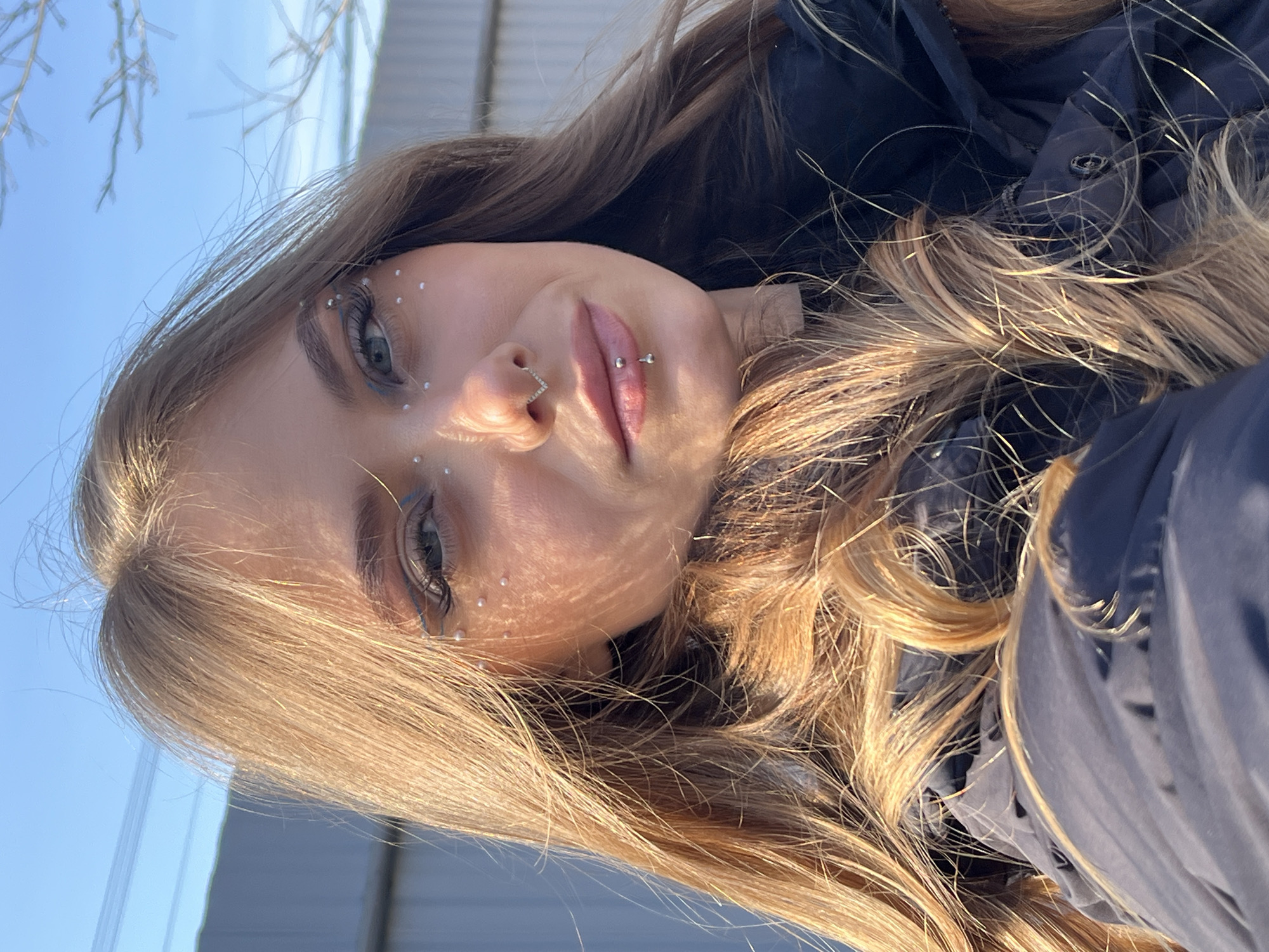 A young woman is depicted outdoors on what appears to be a sunny day. She is wearing a dark-colored jacket and has long, wavy blonde hair. She has noticeable makeup, including eyeliner and small white dots on her face, possibly decorative accents. She also has piercings on her nose and lip. The background shows a glimpse of a blue sky and what seems to be a building or structure with a gray surface. There are bare branches visible in the upper left corner. The location, Фоки, Russia, cannot be verified from the image.