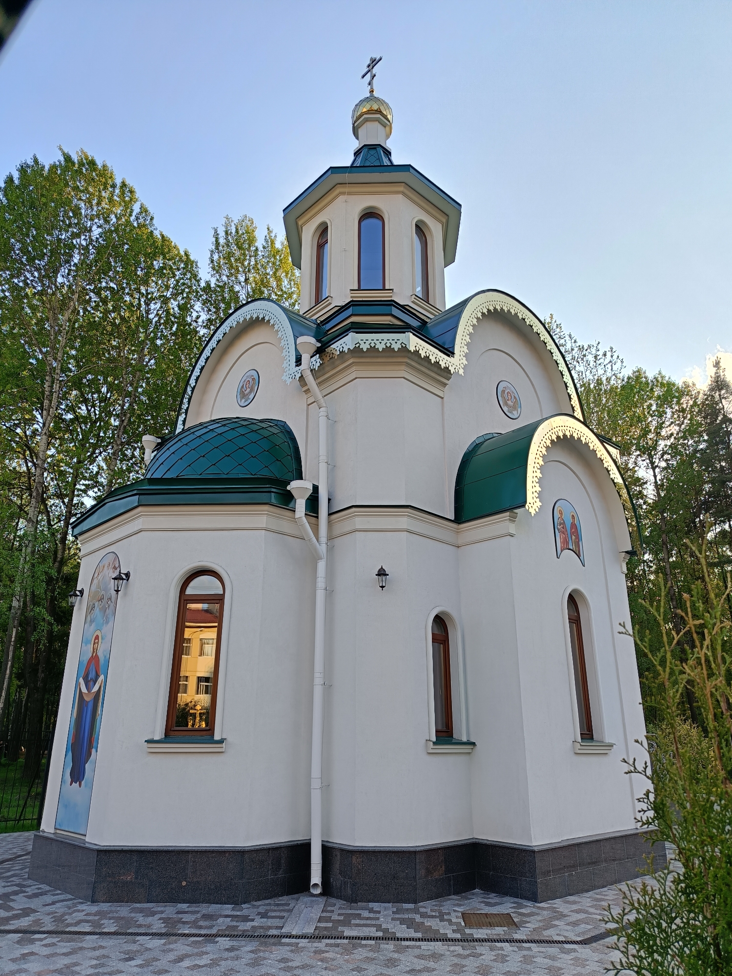 The image depicts a white Eastern Orthodox church, topped with a green roof and a golden dome surmounted by a cross. The building features rounded arch windows with brown trim, decorative painted icons on its walls, and white trim. The church sits on a dark gray foundation, with a paved walkway surrounding it. Green trees partially obscure the view of the church, suggesting a wooded or park-like setting. The sky is blue, indicating a clear day. The location is in Saint Petersburg, Russia.