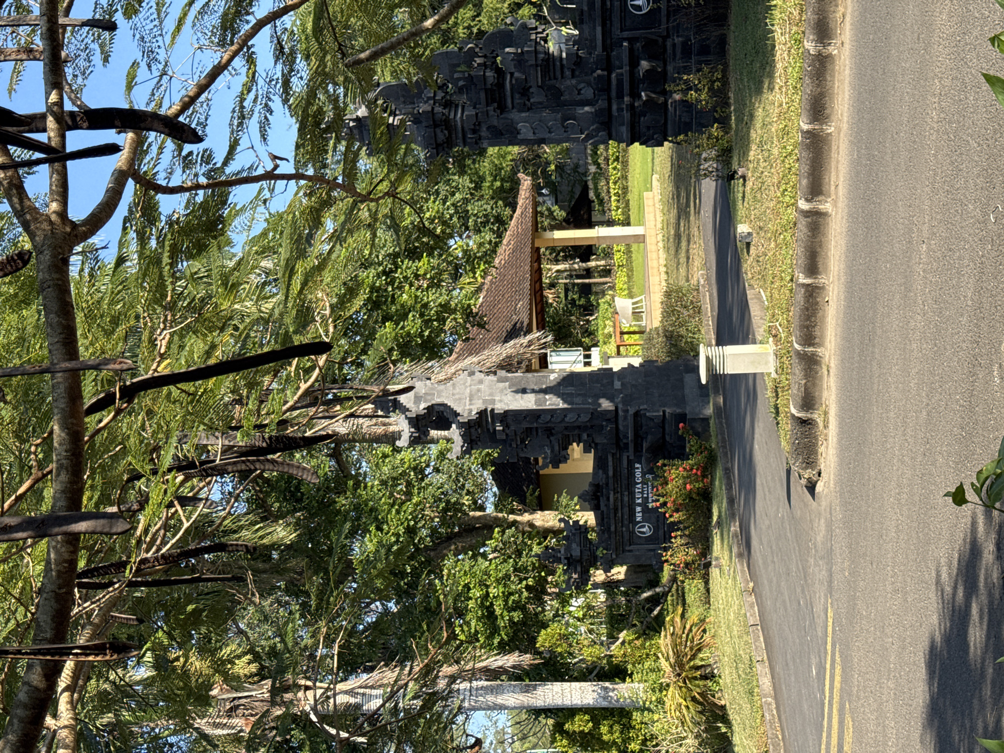 The image depicts the entrance to the New Kuta Golf course in Kuta Selatan, Bali, Indonesia. A black stone Balinese gate frames a view of a small pavilion with a tiled roof, visible through the gate. The foreground features trees with long, dark seed pods hanging from their branches. A paved road runs alongside the entrance, and a sign for New Kuta Golf is visible near the gate. The overall environment appears lush and green.
