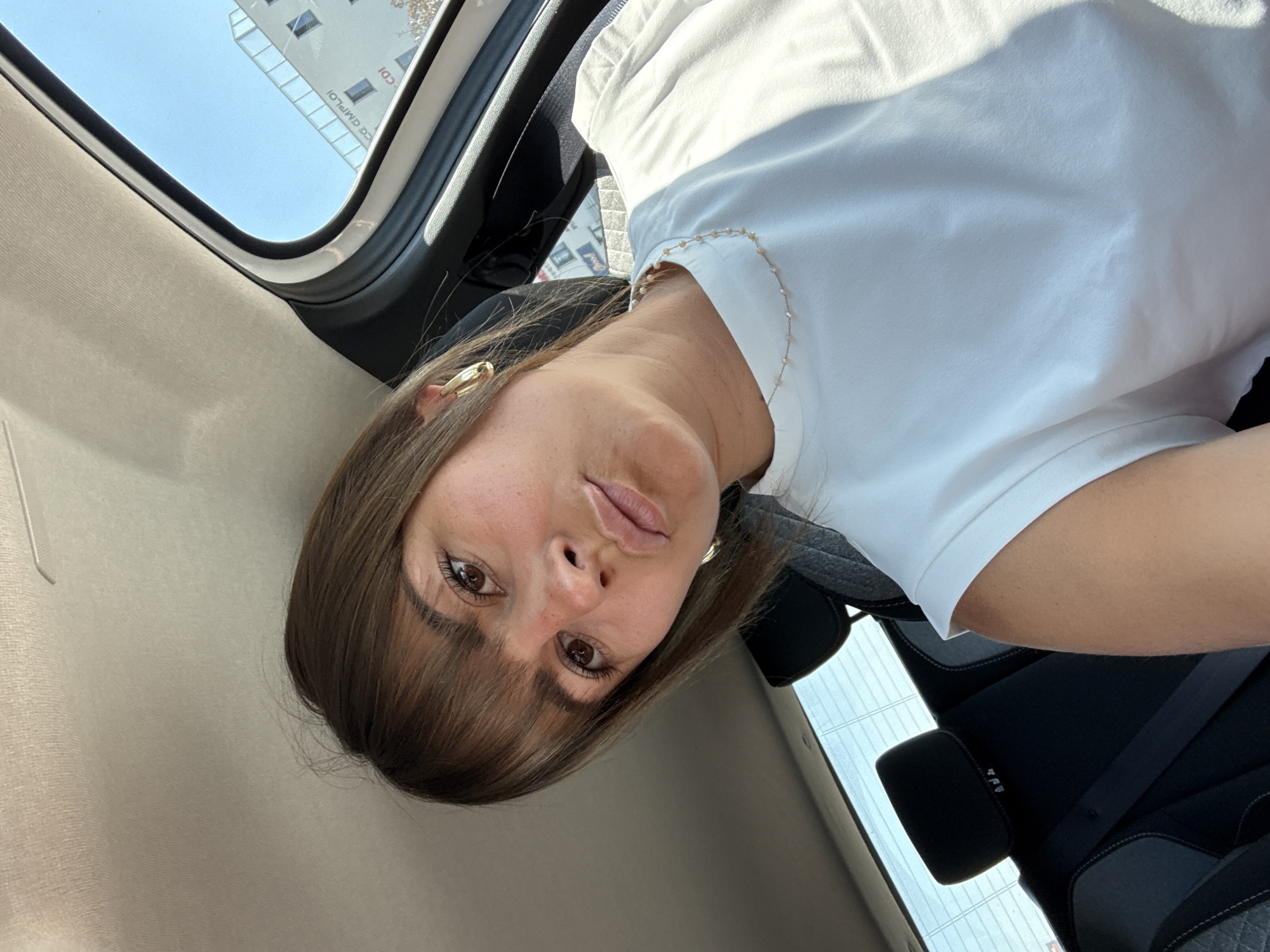 A woman is photographed inside a car, likely in Andrézieux-Bouthéon, France, based on the partial view of a building sign reflecting "CDI". She has brown hair with bangs and is wearing a white t-shirt and a gold necklace with beads. She is looking directly at the camera. The photo is taken at an angle, showing the car's interior including the roof, seat, and seatbelt. The outside sky is visible through the sunroof.