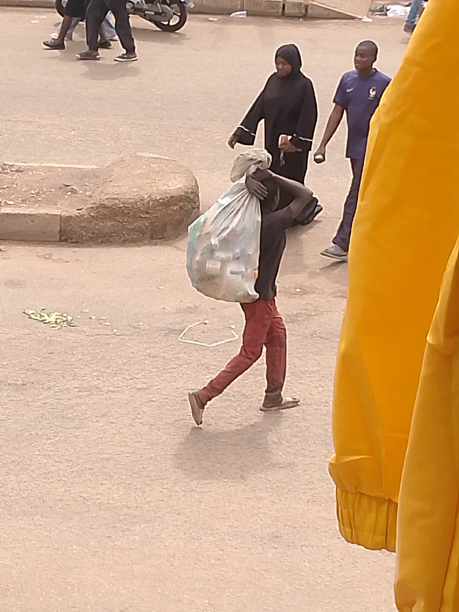 In Bukuru, Nigeria, a person in red trousers and a dark shirt is carrying a large, transparent plastic bag filled with items over their shoulder. They are walking to the right, with their right leg extended. To their left, a woman in a black hijab and long black dress walks with a phone in her hand, and a young man in a blue France soccer jersey walks to her right. In the background, other individuals are visible, and a motorcycle is parked to the left. The ground is paved and appears to be a street or market area. A bright yellow fabric is prominently featured on the right side of the image, obscuring part of the scene.