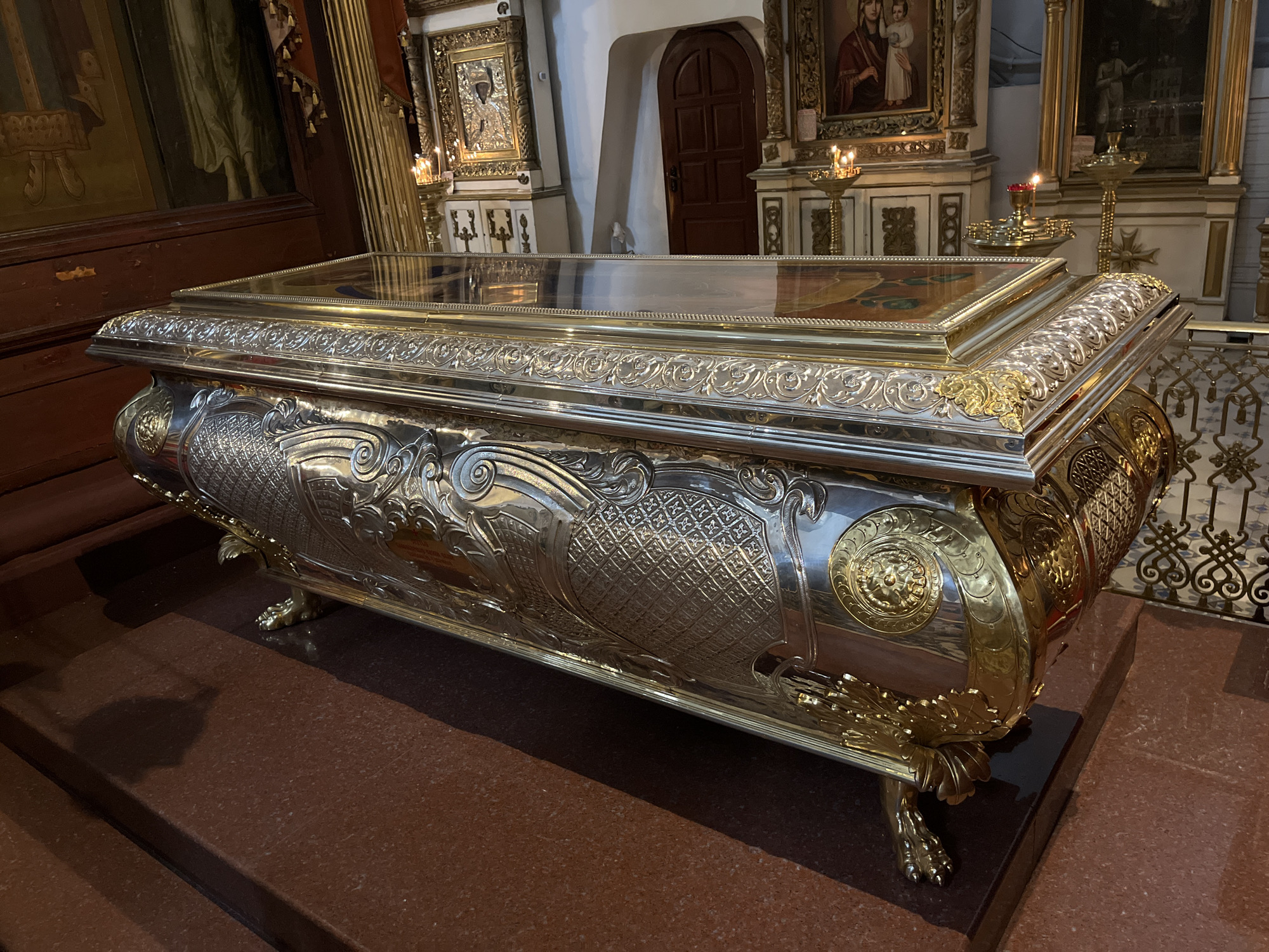 The image shows a richly ornate sarcophagus, likely made of silver and gold, displayed within an Orthodox church. The sarcophagus is rectangular with a glass top, allowing a view of its contents, which appear to be religious relics or icons. The sides of the sarcophagus are elaborately decorated with intricate carvings, embossed patterns, and gilded accents, including floral motifs and scrolling designs. It rests on four decorative golden legs shaped like animal paws. The background reveals the interior of a church, with religious icons, chandeliers, and altar elements visible. The flooring is a dark red, polished stone. The sarcophagus is positioned centrally in the frame, dominating the scene. This sarcophagus is located in the Dormition Cathedral of the Pskov Kremlin in Pskov, Russia.