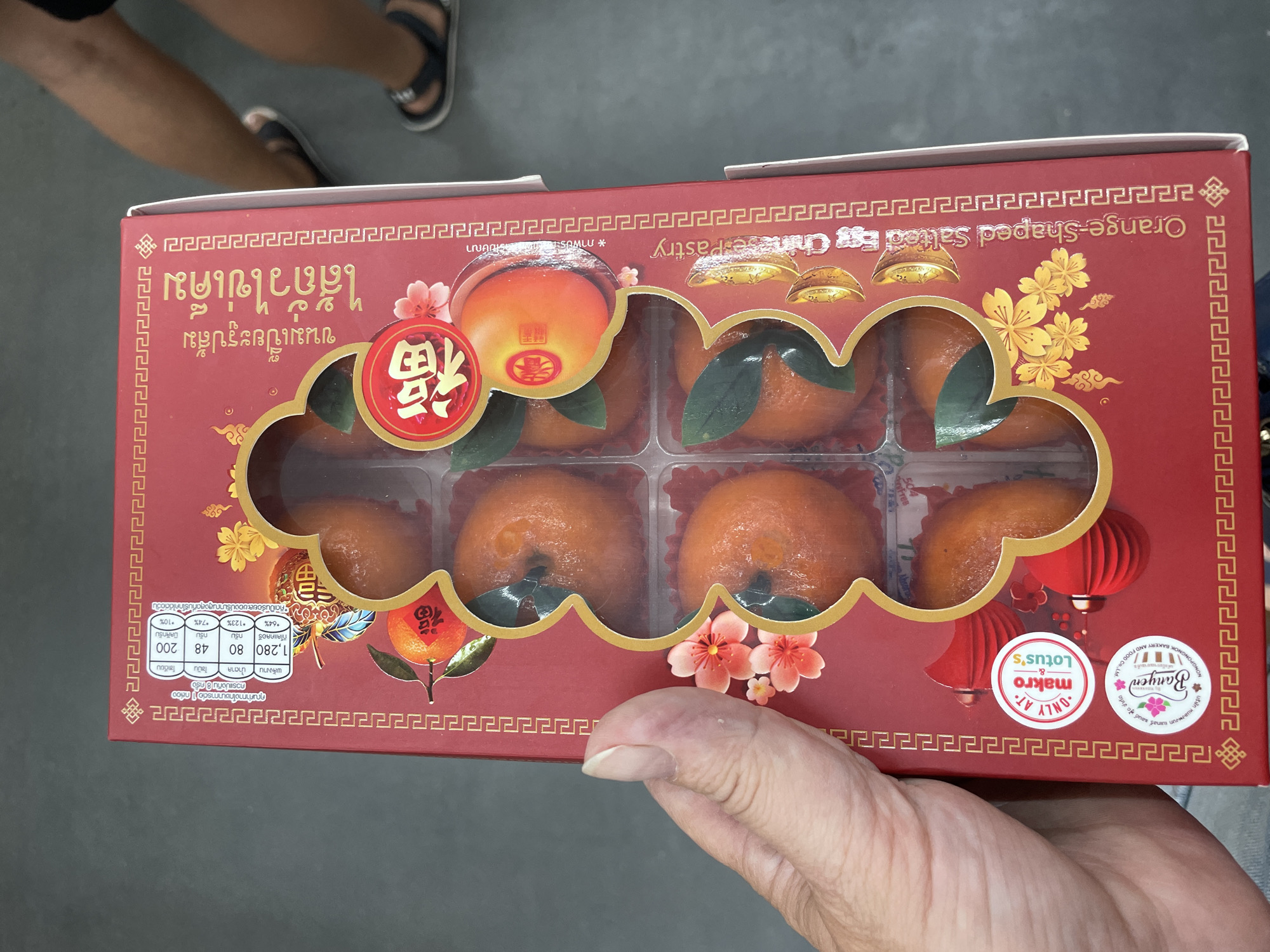 The image shows a close-up of a red gift box of orange-shaped pastries, likely for a holiday celebration such as Chinese New Year. The box is decorated with traditional Chinese motifs, including lanterns and floral designs, and features Thai text. The pastries are visible through a clear plastic window in the center of the box, arranged in two rows. Below the window, there are two circular stickers, one with "Only at makro & Lotus's" and another with "Banyen". The text on the box indicates the product is "Orange-Shaped Salted Egg Chinese Pastry". In the background, the legs and feet of two individuals wearing sandals are visible, suggesting an indoor setting, possibly a market or store. The overall scene is consistent with a retail environment in Pattaya City, Thailand, where such festive products are commonly sold.