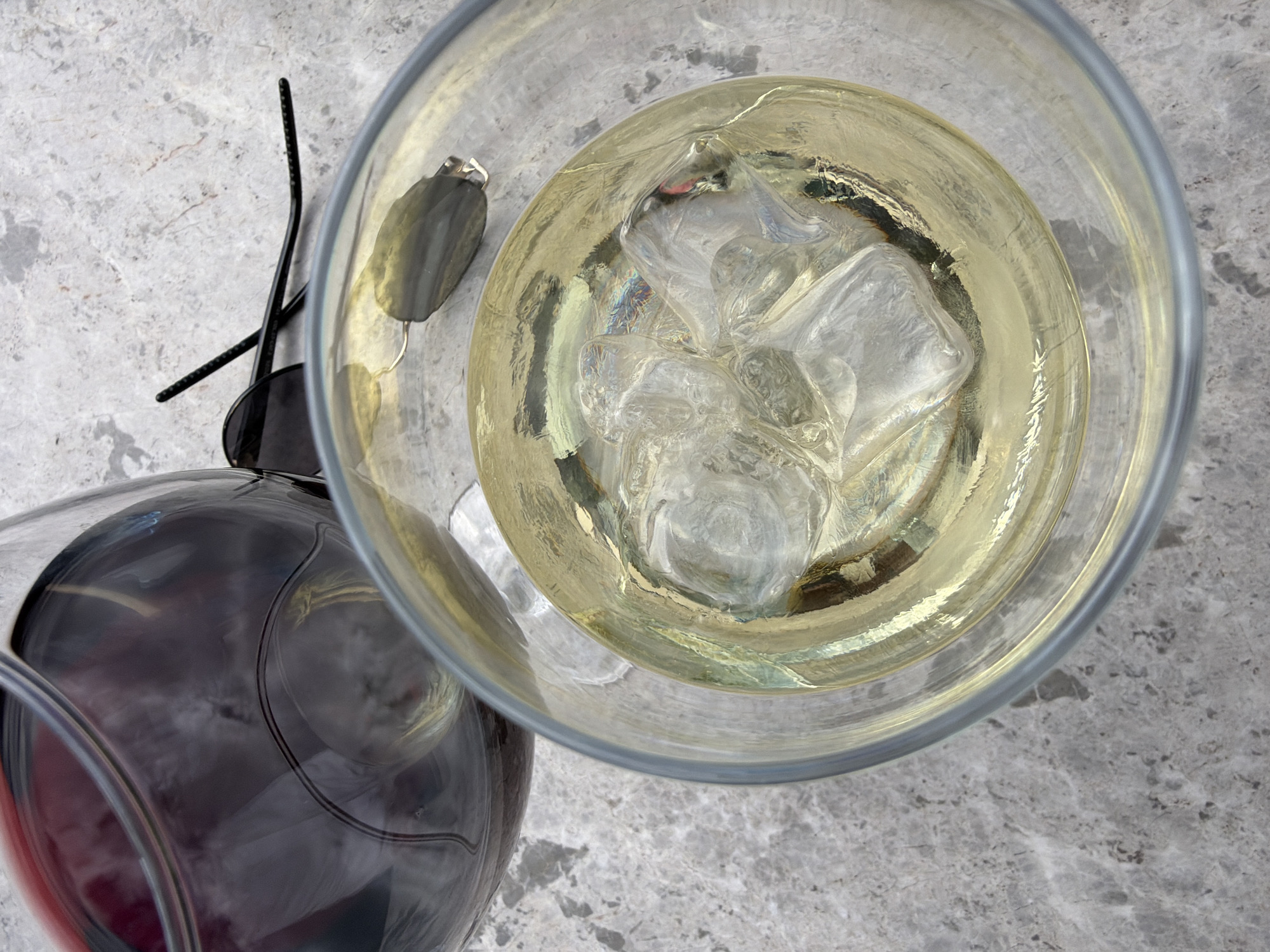 An overhead shot displays a beverage with ice in a clear glass and sunglasses on a gray, textured surface. The beverage glass, filled with a light-colored liquid and ice, is positioned slightly off-center towards the upper right. Next to it, on the left side of the frame, are dark-colored sunglasses with a visible lens and frame. The glasses are partially within another transparent container. The surface exhibits a pattern with shades of gray, resembling a stone or composite material. The location is unknown.