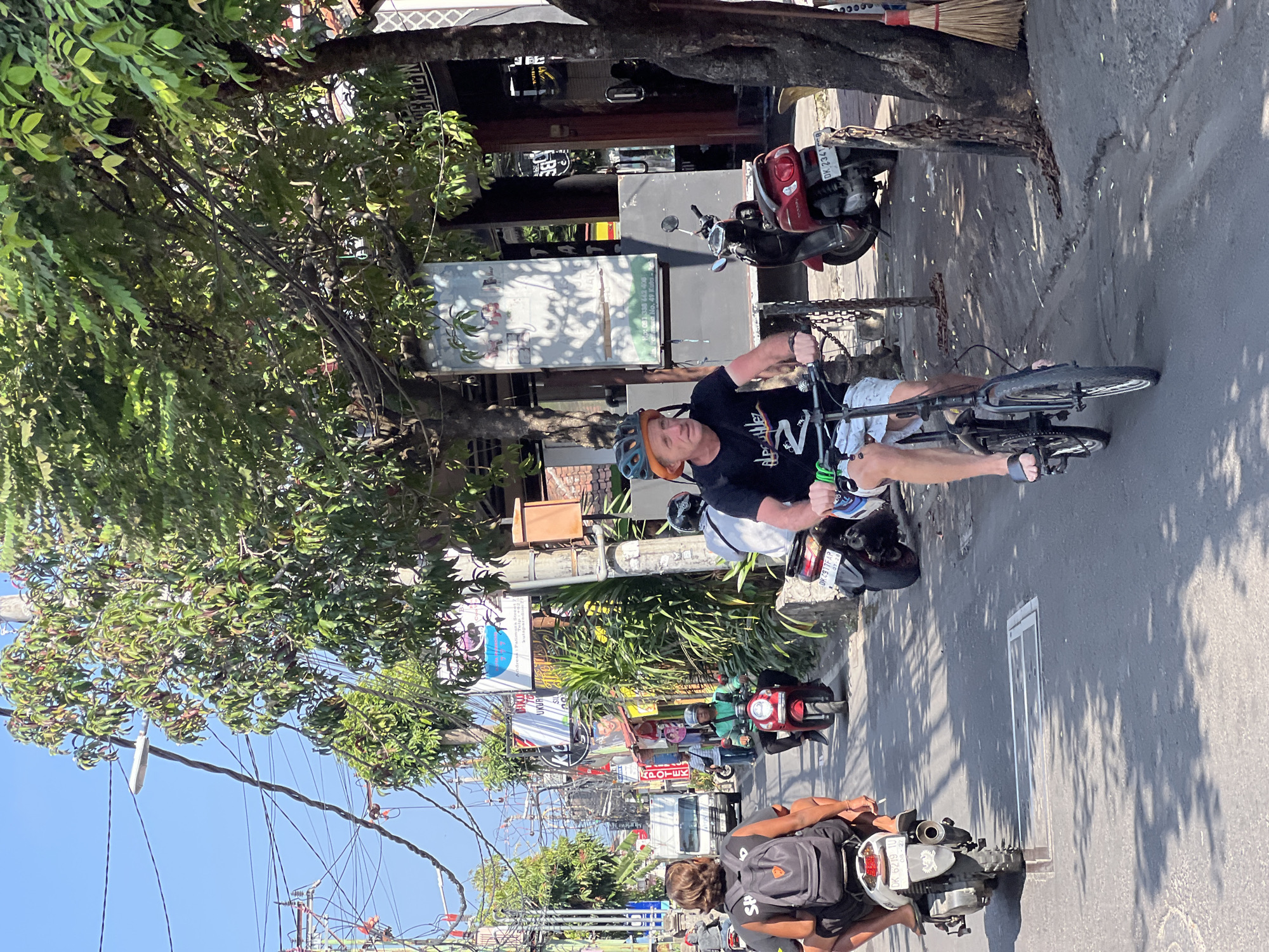 In Kuta, Indonesia, a sunny street scene unfolds with several people on motorcycles and bicycles. In the foreground, a man wearing a helmet and shorts rides a black folding bicycle. Behind him, a person with a backpack rides a white scooter, followed by another person on a red scooter. Further down the street, a man in a green shirt rides a red scooter, and a white van is visible. The street is lined with trees and buildings displaying various signs, including what appears to be a tattoo parlor and shops. Overhead, a complex network of electrical wires crisscrosses the sky, and a street lamp is visible. The road surface is asphalt, with some cracks and shadows cast by the trees.