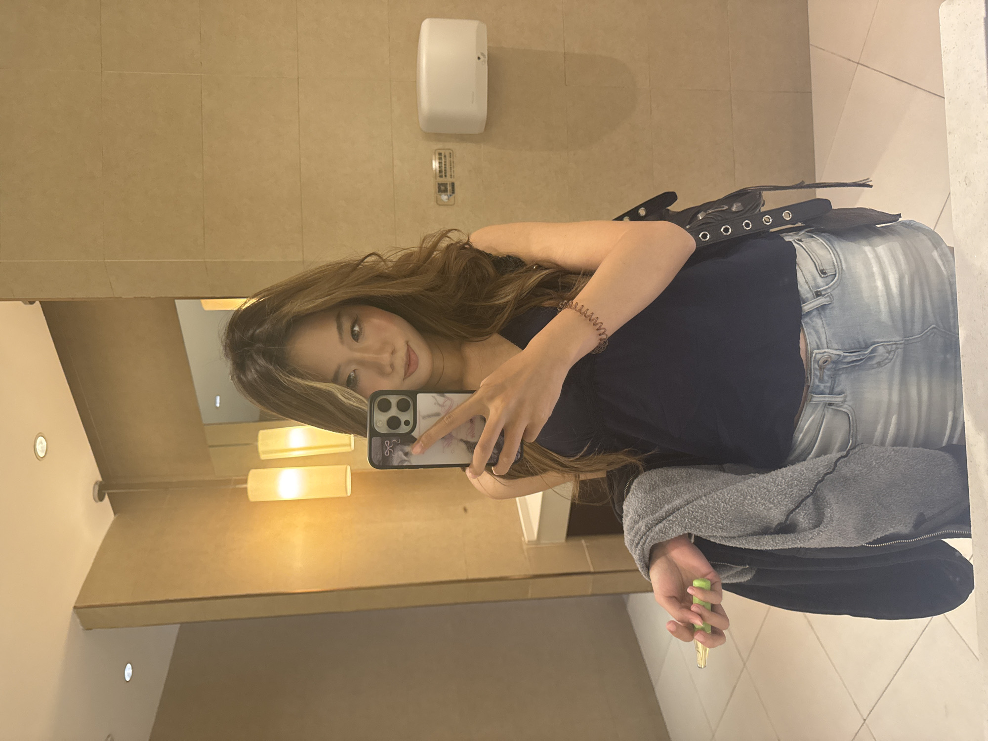 A young woman stands in a restroom, likely in Yuzhong District, China, based on the photo's origin. She holds an iPhone in her left hand, taking a selfie in the mirror. She is wearing a dark blue top, light blue jeans, and a gray zip-up jacket. Her hair is long and light brown. A black bag hangs from her shoulder. In her right hand, she holds a green and gold object. The restroom features beige tiled walls, a white hand dryer, and bright overhead lighting.