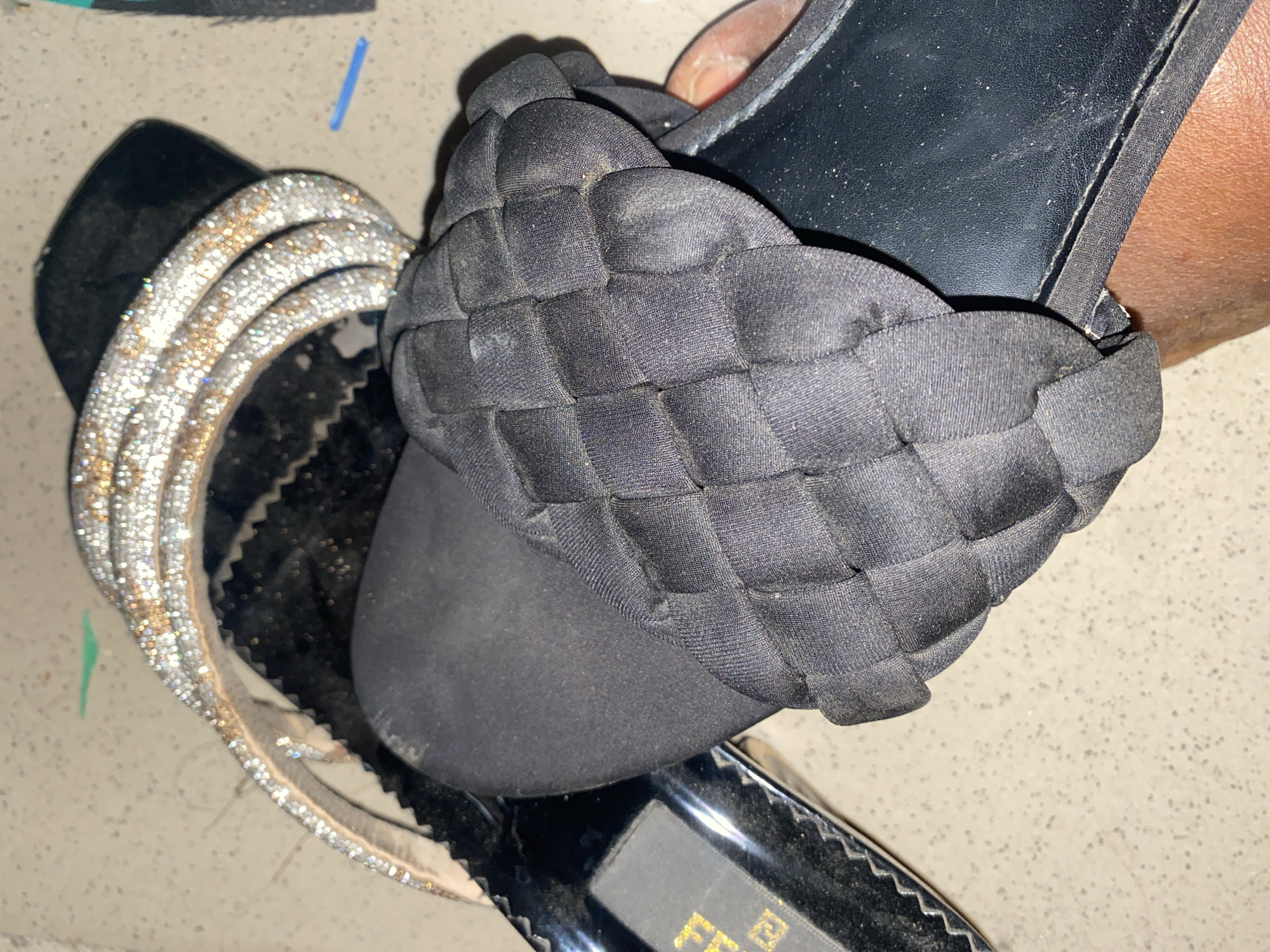 The image shows a close-up, overhead view of a black slide sandal with a woven fabric strap across the foot. The sandal is on a speckled light-colored surface, possibly a countertop or floor. To the left of the sandal, partially visible, is another sandal with a black sole and a strap embellished with multiple rows of sparkling rhinestones. A portion of a person's skin, appearing to be a hand or arm, is visible at the bottom right of the frame, holding the sandal. The overall lighting suggests an indoor setting. The image does not contain any identifiable landmarks or elements that would suggest its location in Jauro Yinu, Nigeria.