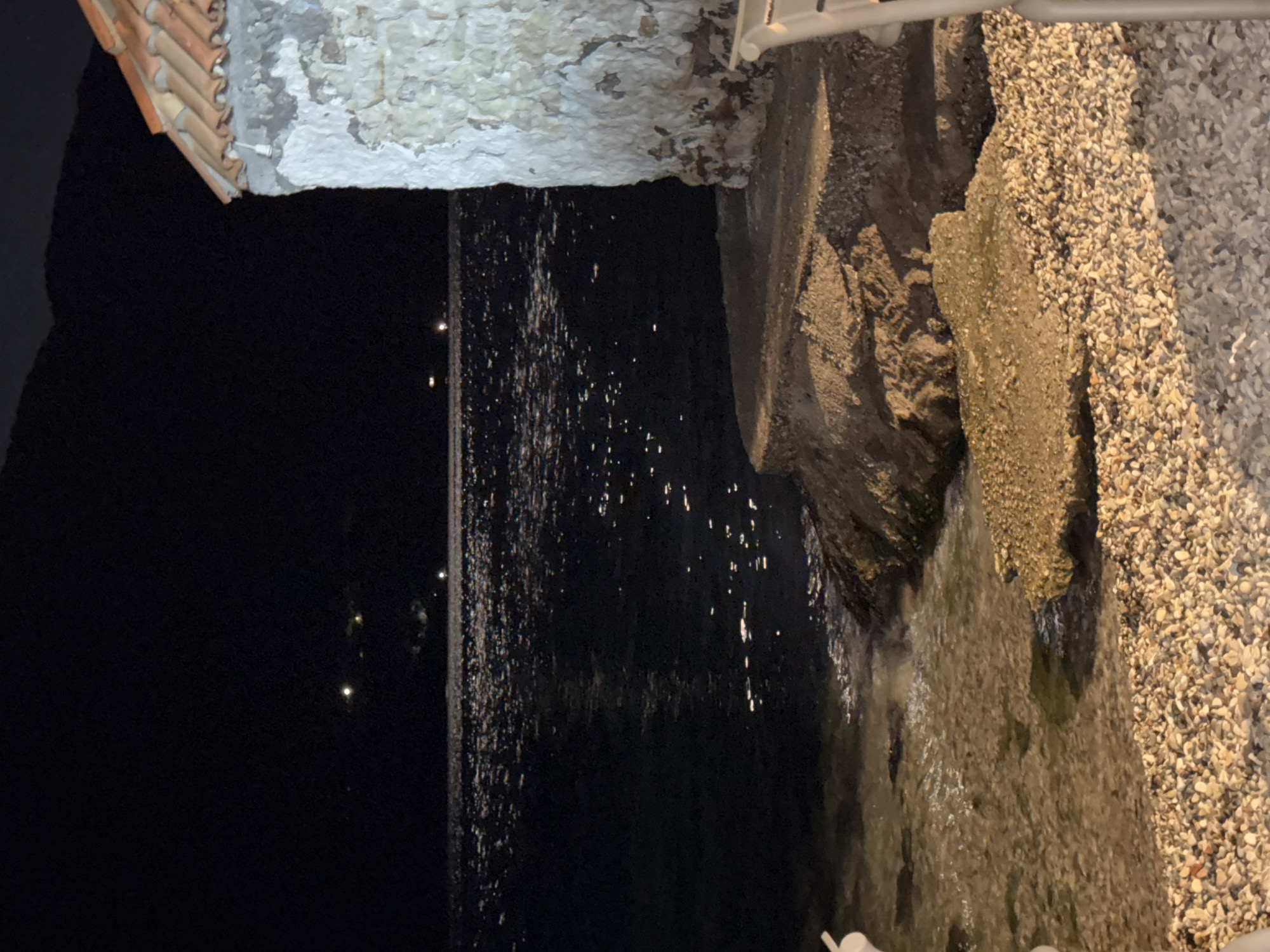 At night, a view of the sea is captured from a pebble beach. The water reflects scattered lights from a distant shoreline, creating a shimmering effect. To the right, a portion of a stone building with a tiled roof is visible. The building's rough texture and weathered appearance are evident. The foreground is dominated by the beach, covered in small, smooth pebbles. The background features a dark, mountainous silhouette against the night sky. The location is the Municipal Unit of Alyzia, Greece.