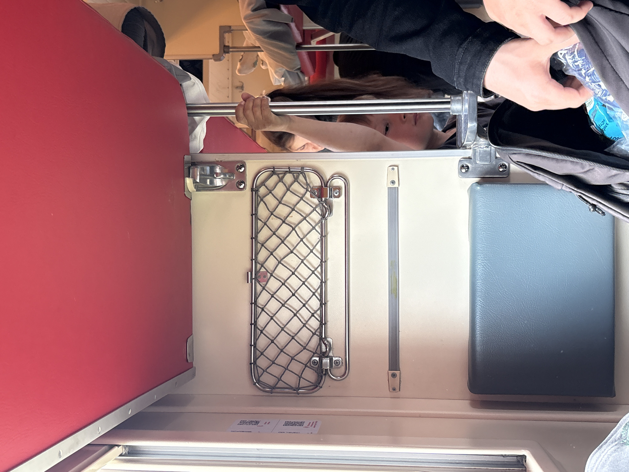This is a medium-shot, indoor, eye-level, slightly-low-angle photograph taken inside a train compartment. The compartment appears to be a typical Russian train carriage, suggesting the location is within Saint Petersburg, Russia, or on a train route originating from or traveling to the city. The focus is on the interior of the train, with a prominent red table and a metal support pole in the foreground.
On the left side of the image, there's a window with a view of the exterior, though it's mostly obscured. Adjacent to the window are two QR code stickers affixed to the wall. Below these stickers, a wire mesh compartment is mounted on the wall. Further down, a pale blue padded seat or cushion is visible.
In the center-right of the frame, a young person, likely a child, is peeking from behind the support pole, looking downwards. Their face is partially visible, showing their brow and eyes. They are holding onto the metal pole with their right hand. Another person, whose hand is visible in the lower right corner, is reaching into a dark backpack. This backpack is placed on the floor of the compartment, and it appears to contain a blue plastic bottle or bag.
The overall color palette is dominated by the vibrant red of the table, the neutral tones of the walls and seats, and the darker colors of the backpack and clothing. The lighting suggests it's daytime, possibly with natural light coming from the window. The scene conveys a sense of travel and everyday life within a confined space.