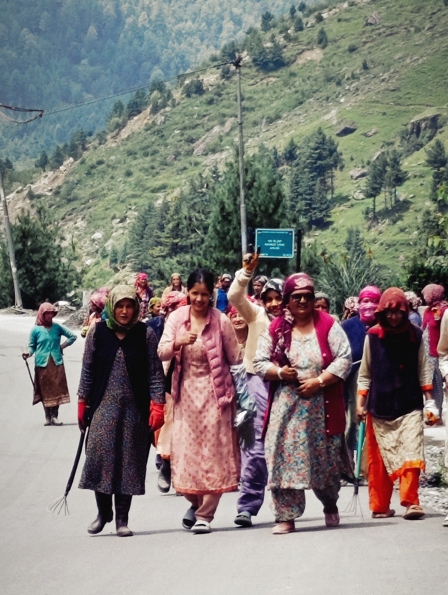 A group of women are walking down a road in Kothi, India. The road is paved and bordered by a steep, green hillside covered in trees and rocks. The women are dressed in a variety of colorful clothing, including scarves, long dresses, and vests. Some of the women are carrying tools, such as a rake and a stick. The weather appears to be sunny and warm. The overall scene suggests a community outing or a procession.