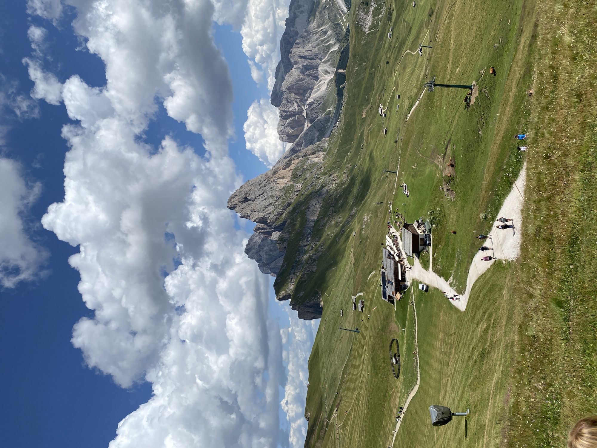 This is a medium shot taken from a high vantage point, looking down on a mountainous landscape. The sky is a vibrant blue, dotted with large, puffy white clouds. The foreground is a lush green meadow, with a winding dirt path leading into the distance. Several people are walking along the path.
Further into the scene, a cluster of buildings with rustic architecture sits on a hillside. These appear to be part of a mountain resort or restaurant, with outdoor seating visible. A ski lift structure is prominent near the buildings.
The middle ground features rolling green hills, some with terraced patterns suggestive of mowing or grazing. More paths and scattered small buildings are visible. Several cows are grazing in a pasture.
The background is dominated by dramatic, jagged mountain peaks, characteristic of the Dolomites. These mountains are steep and rocky, with patches of green vegetation on their lower slopes. The lighting suggests it is daytime, with the sun casting shadows that highlight the contours of the landscape. The overall impression is a scenic mountain vista in a resort area.
The image appears to be taken in the Dolomites region of Italy, possibly near a popular hiking or skiing destination.