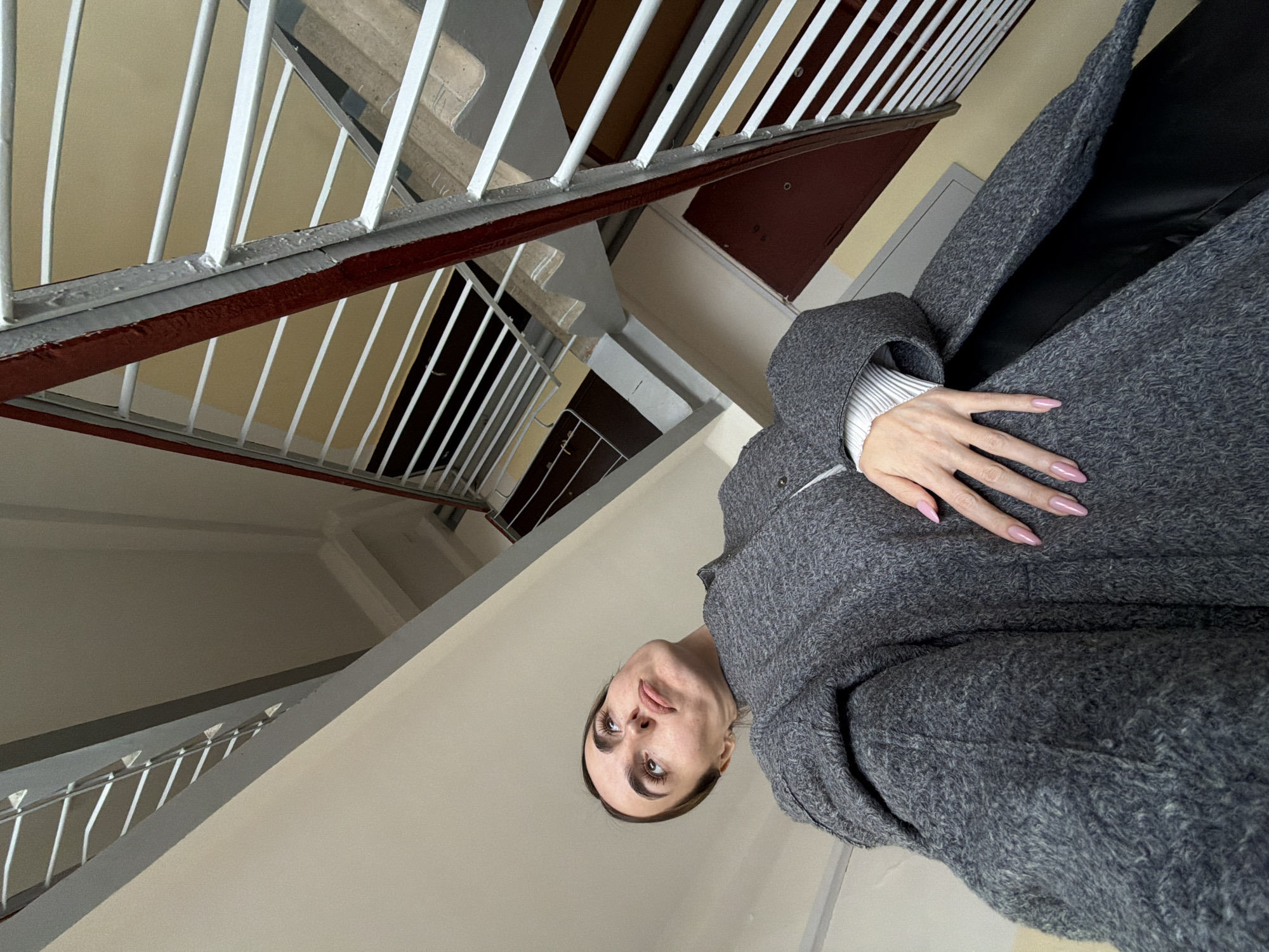 A medium, tilted, indoor shot presents a woman in a grey wool coat with long pink nails, set against the backdrop of an old staircase. The staircase features white bars and brown rails. The walls are painted in light beige. This scene is likely in the city of Izhevsk, Russia.