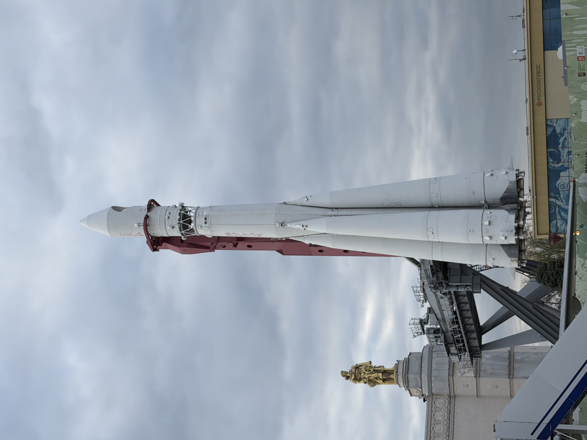 Here is a description of the image:
The image shows a Soyuz rocket on display at the VDNH (Exhibition of Achievements of National Economy) in Moscow, Russia. The rocket is white with red accents and Cyrillic text. It is positioned horizontally on a gantry. Behind it is a cloudy sky. A statue of a man holding a steering wheel sits on top of a building below the rocket. A building with signage is to the side of the rocket.