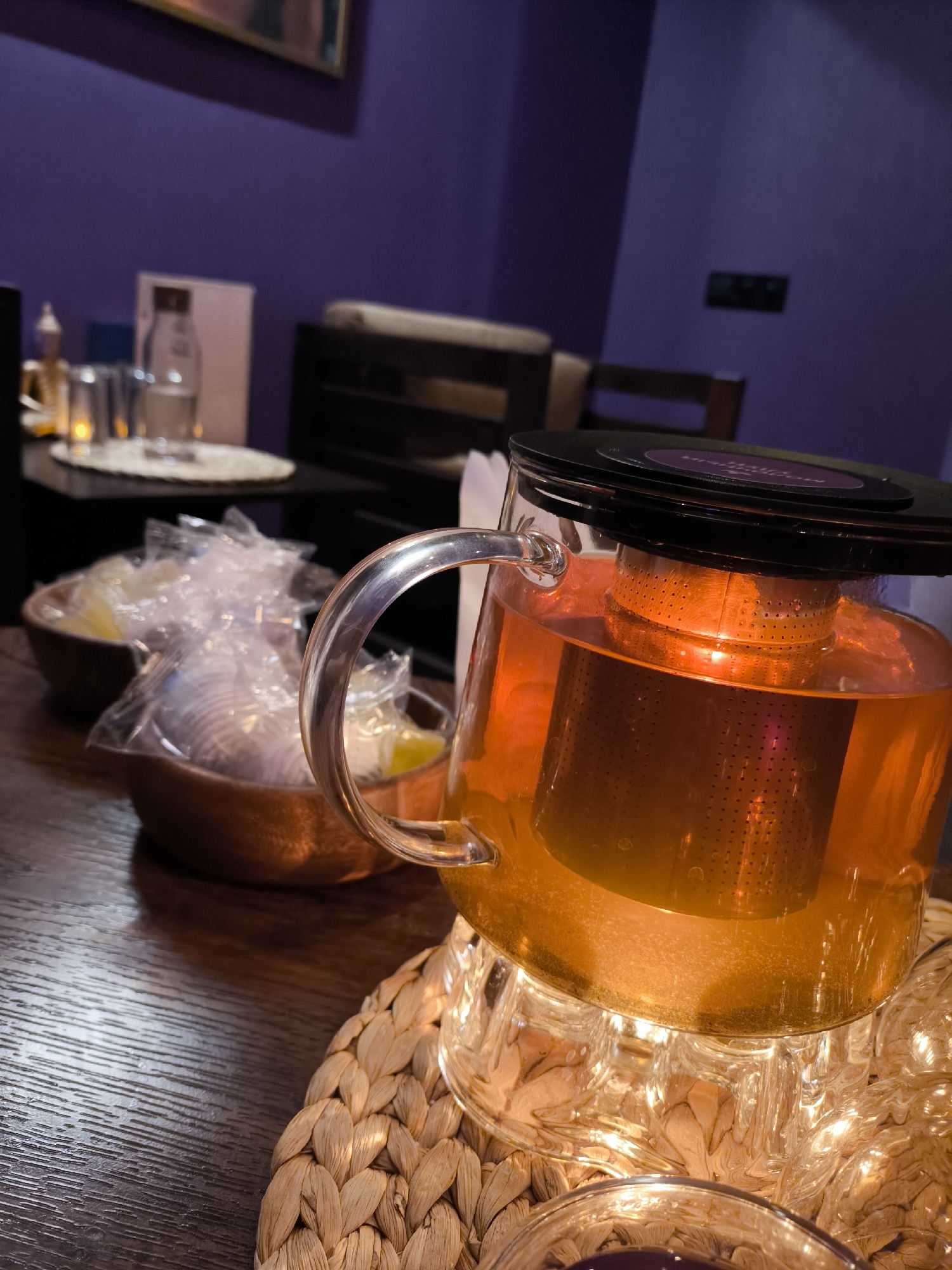 Here is a detailed description of the image:
**Scene:** The image captures an indoor setting, likely a tea room or lounge, in the city of Khimki, Russia. The room has a sophisticated and cozy ambiance with purple walls.
**Objects:**
* **Tea Pot:** The prominent subject is a clear glass teapot filled with amber-colored tea. It has a built-in metal infuser and a black lid.
* **Tea Warmer:** The teapot rests on a small, decorative tea warmer made of clear glass and lit with small lights, creating a gentle glow. It sits on a woven placemat.
* **Tea Cup:** A glass tea cup is partly visible near the bottom edge of the image.
* **Bowls:** In the background, there are two wooden bowls containing individually wrapped items (possibly lemon slices or sugar cubes).
* **Background Items:** Other items are visible in the background, including glasses, a small decorative golden statue or ornament, a menu, and dark furniture.
**People:** The image does not contain any people.
**Actions:** There are no apparent actions, as the scene is static. The focus is on the prepared tea and its presentation.
**Interactions:** There is no direct interaction between objects or people in the scene.
**Environment:** The lighting in the room is warm and subdued, enhancing the cozy and relaxing atmosphere. The purple walls contribute to the overall sophisticated feel.