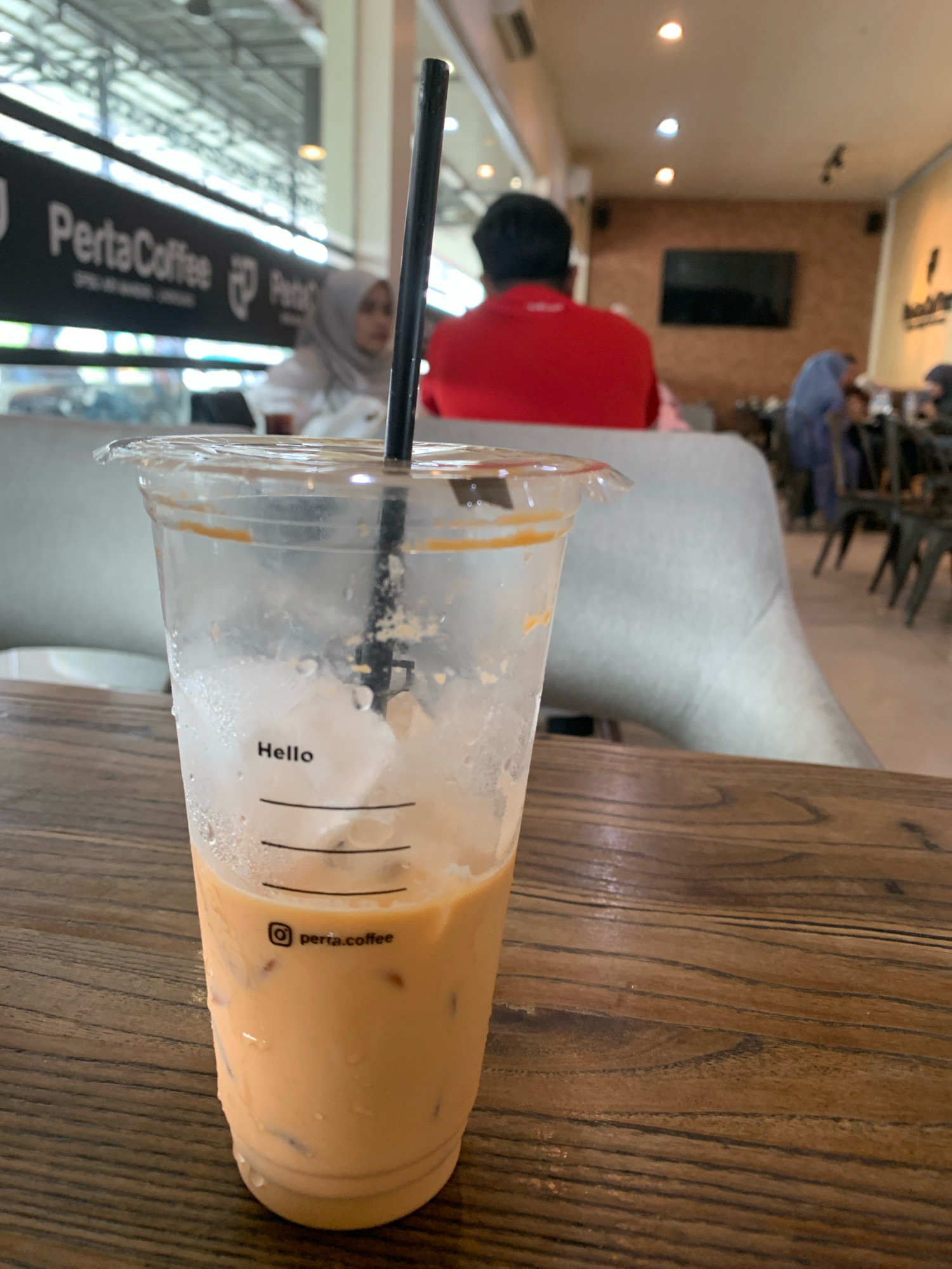 An iced beverage in a clear plastic cup with a black straw sits on a wooden table in the foreground of an indoor cafe. The cup has ice cubes and a pale orange liquid. Text on the cup reads "Hello" and "perta.coffee". In the background, several people are visible, including a person wearing a red shirt and another wearing a grey hijab. A sign with "PertaCoffee" is mounted on the wall behind them. The environment appears to be a coffee shop in Meunasahceubrek, Indonesia.