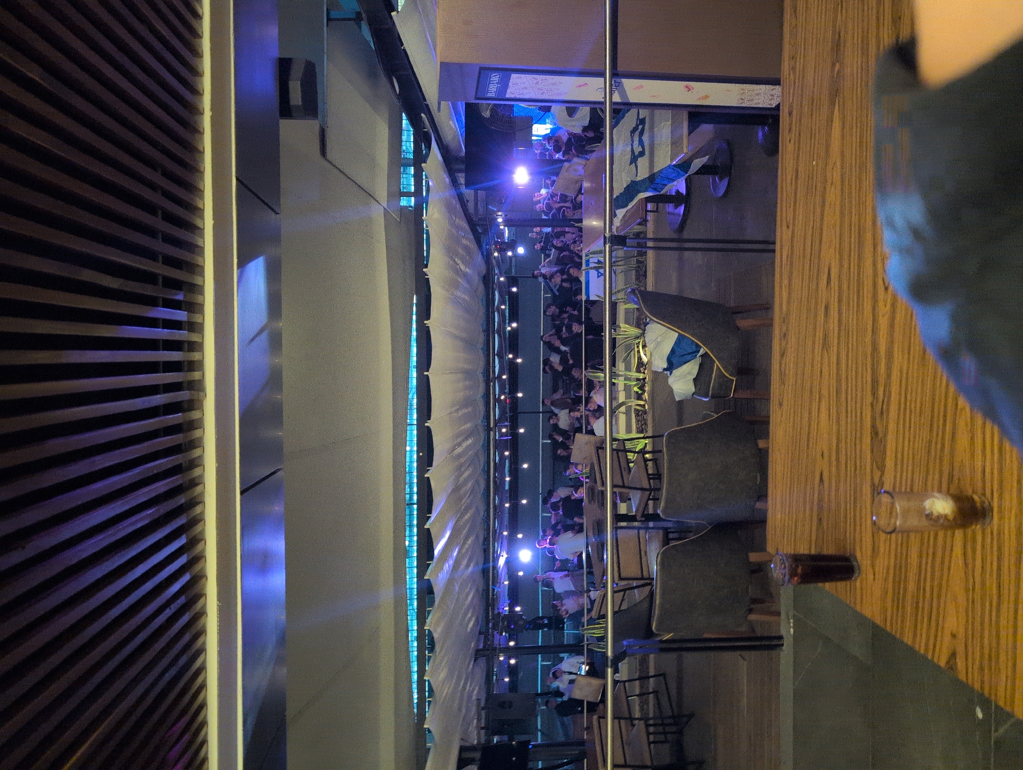 The image depicts an outdoor seating area at a restaurant named Barrabas, located in Mexico City, Mexico. The scene is set under a canopy with blue lights. Numerous people are seated at tables, suggesting an event or gathering. Several chairs and tables are visible, along with glasses on the table. A wooden panel flanks one side of the view. The overall atmosphere suggests an evening social gathering.