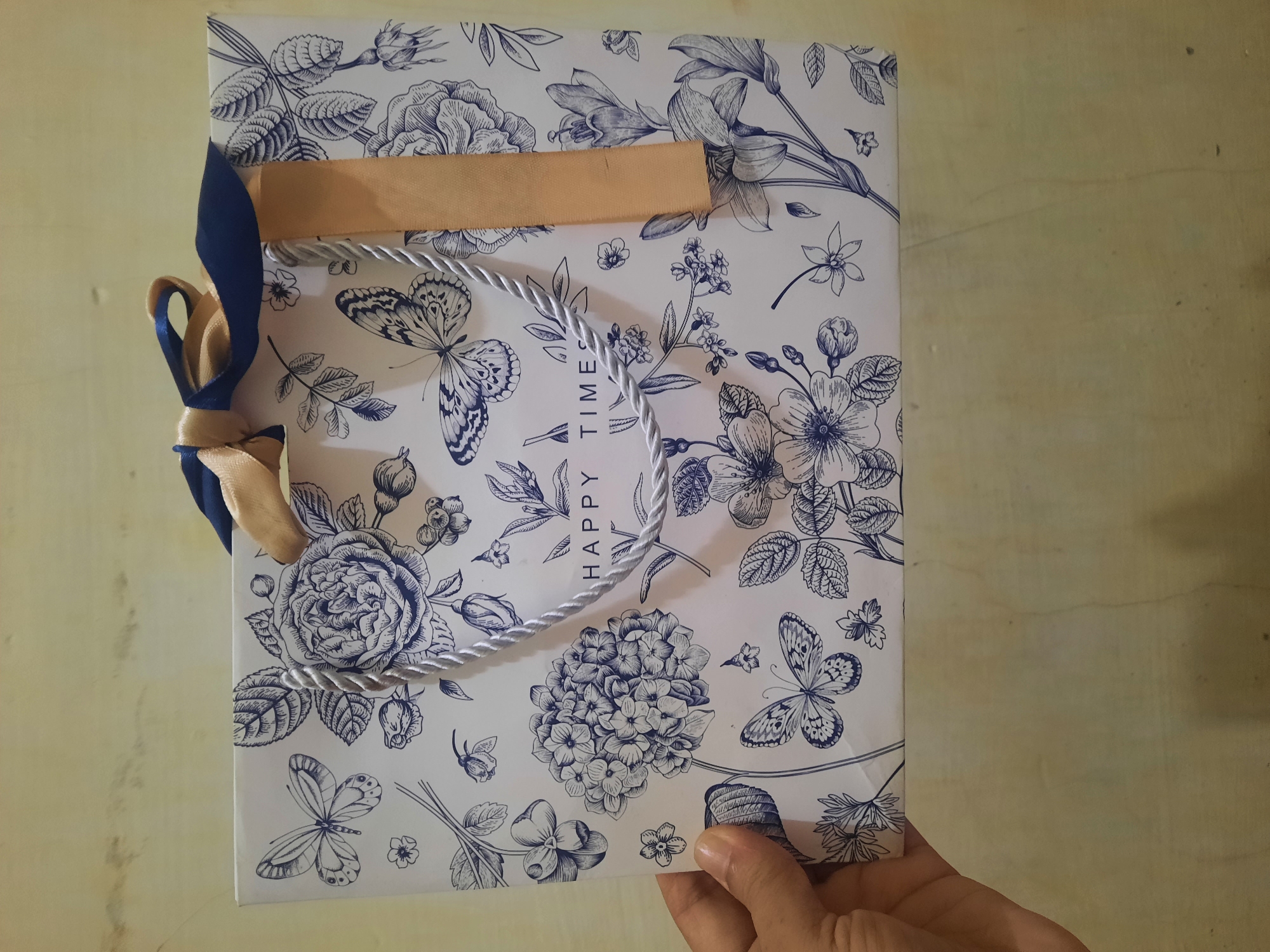 A close-up, eye-level shot shows a white gift bag with blue floral and butterfly illustrations. A person's hand is visible on the left side of the image, holding the bag. The bag has rope handles and is adorned with a blue and gold ribbon tied in a bow at the top. The text "HAPPY TIMES" is printed in the center of the bag. The background is a plain, light-colored wall. This image was captured in Lhokseumawe, Indonesia.