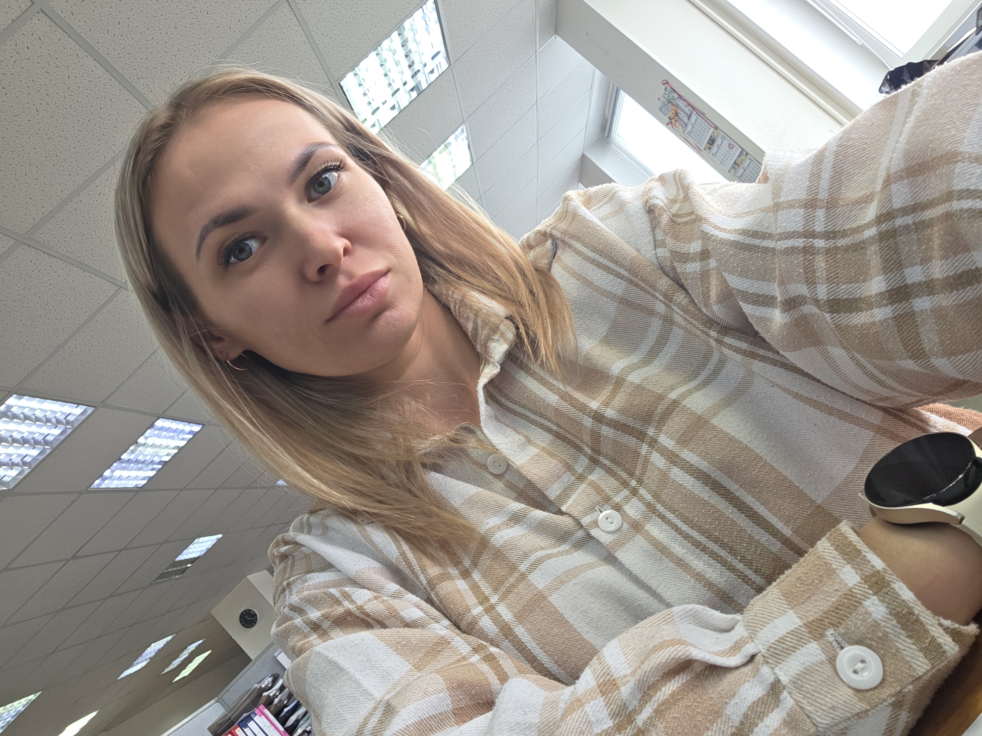 A woman with blonde hair is taking a selfie in an indoor location. She is wearing a beige and white plaid shirt. Her left wrist is visible, with a smartwatch. The background consists of a ceiling with fluorescent lights and what appears to be an office environment with a window, a calendar, and possibly filing cabinets. The location appears to be an office building in a city, however, no explicit connection can be made to Chaikovsky, Russia from the image.