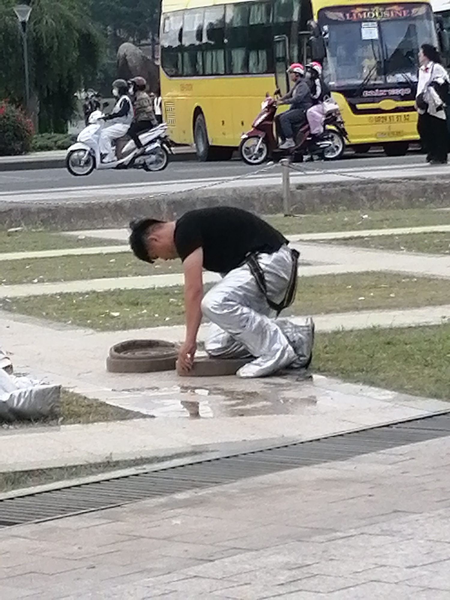 A man in a black shirt and shiny silver pants is kneeling on a paved area, reaching towards a circular metal manhole cover. He is positioned in front of a grassy area with some pathways and a large metal grate. In the background, a yellow bus with "LIMOUSINE" written on it is parked, and two people are riding a white scooter with another scooter nearby. A woman in a white shirt and dark pants is standing to the right of the bus. The scene appears to be in Da Lat, Vietnam, as indicated by the presence of a limousine bus, commonly used for transportation in the city, and the overall setting resembling an urban public space.