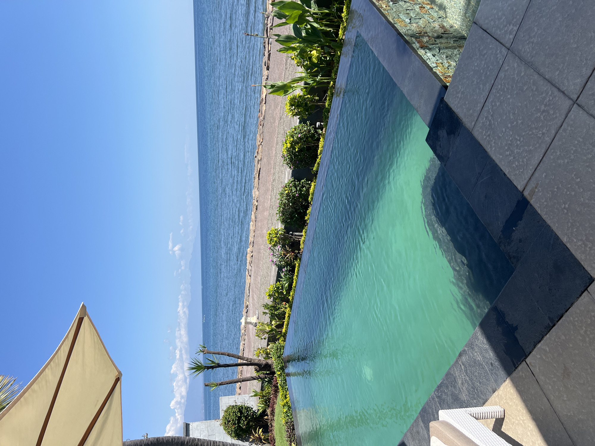 An infinity swimming pool with turquoise water is in the foreground, bordered by dark stone coping and steps. Beyond the pool is a landscaped area with lush green plants and some small trees. A stone pathway runs along the edge of the landscaping, separating it from a body of blue water, likely the sea. In the distance, hazy mountains are visible against a clear blue sky. The scene suggests a tranquil, resort-like setting, possibly in Buleleng, Indonesia, given the tropical vegetation and coastal view.