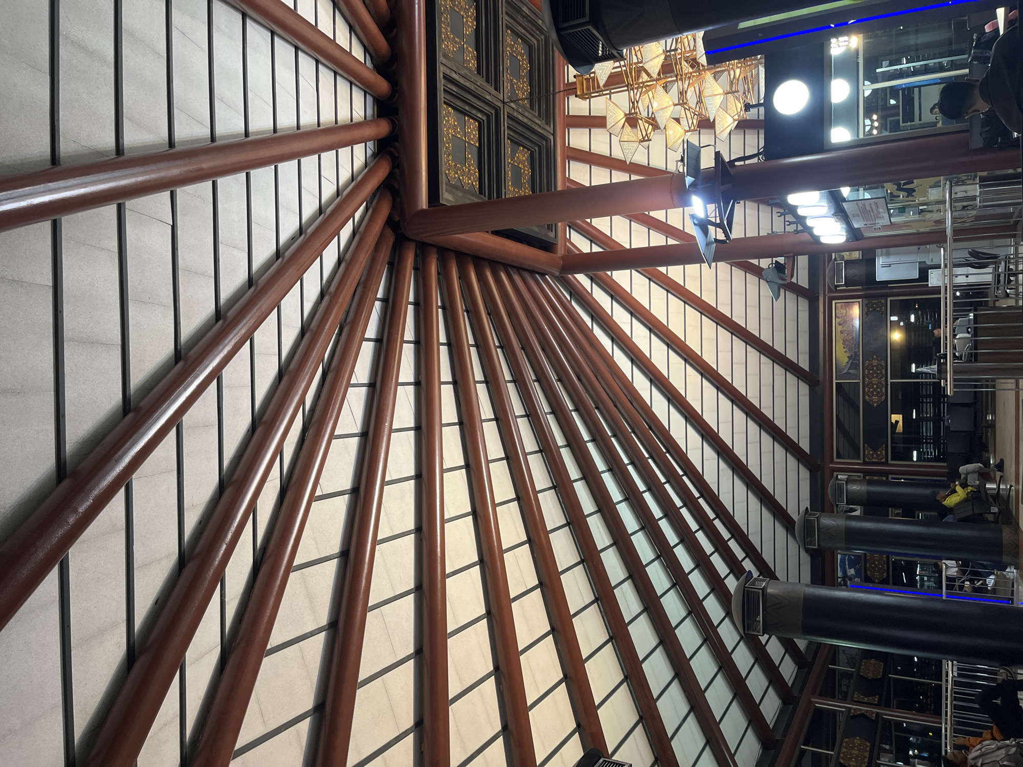 The image depicts the interior of a building in Tangerang, Indonesia, showcasing a radial pattern of wooden beams extending from a central point towards the perimeter. The beams are dark brown, contrasting with the lighter-colored panels that fill the spaces between them. The central structure from which the beams emanate is decorated with gold-colored ornaments. Overhead, there is a chandelier and modern lighting fixtures. The reflection of a staircase is visible on the right side of the image. Air conditioning units are visible on the right as well. The background shows people standing or sitting, but they are indistinct due to the focus on the architectural details. The flooring appears to be comprised of light-colored rectangular tiles.