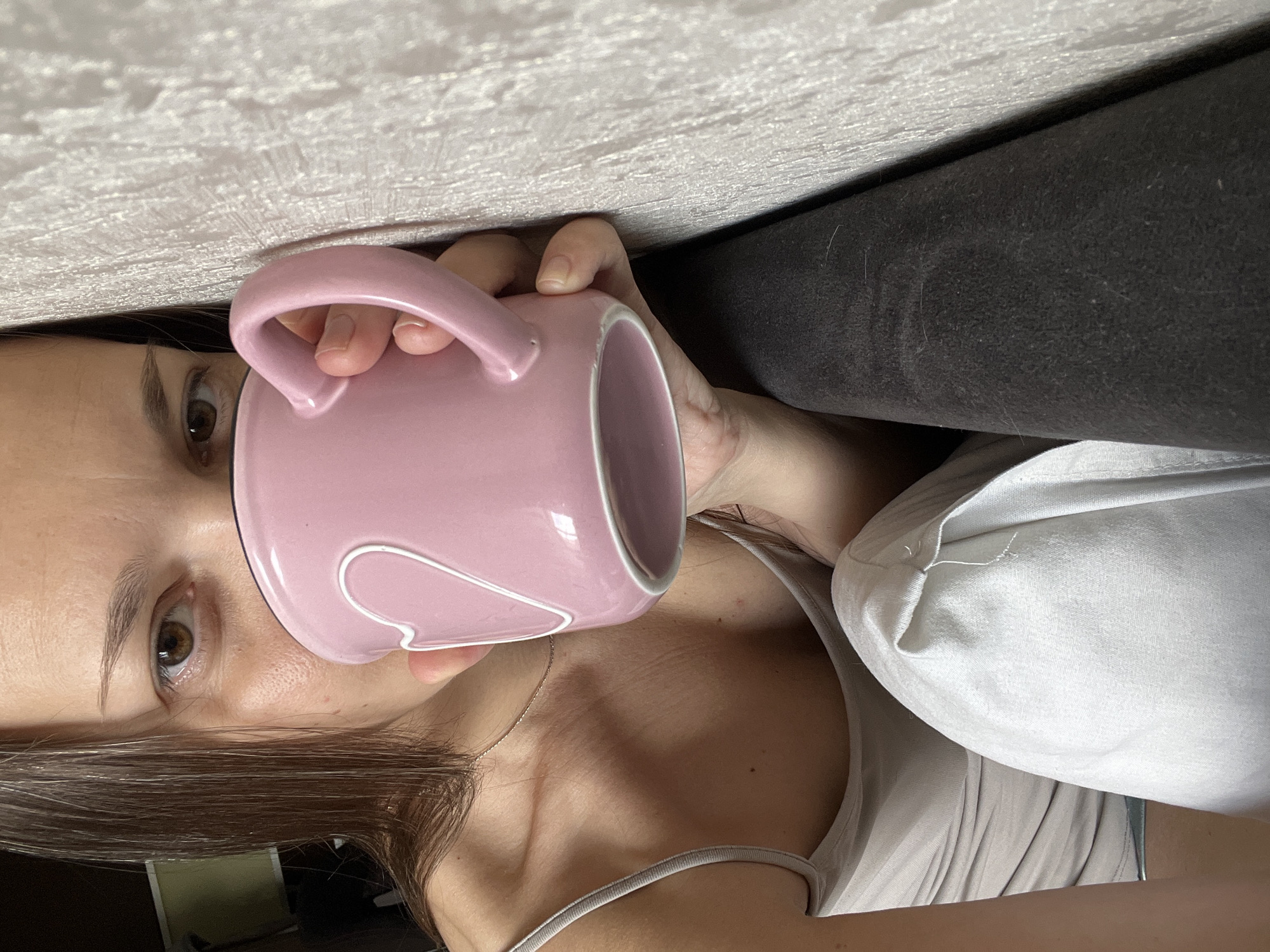 A woman with dark hair is holding a pink mug with a white heart design to her lips. She is wearing a light-colored camisole and light-colored pants. She is leaning against a textured wall. The setting is indoors, and it is not possible to determine the specific location in Tolyatti, Russia.