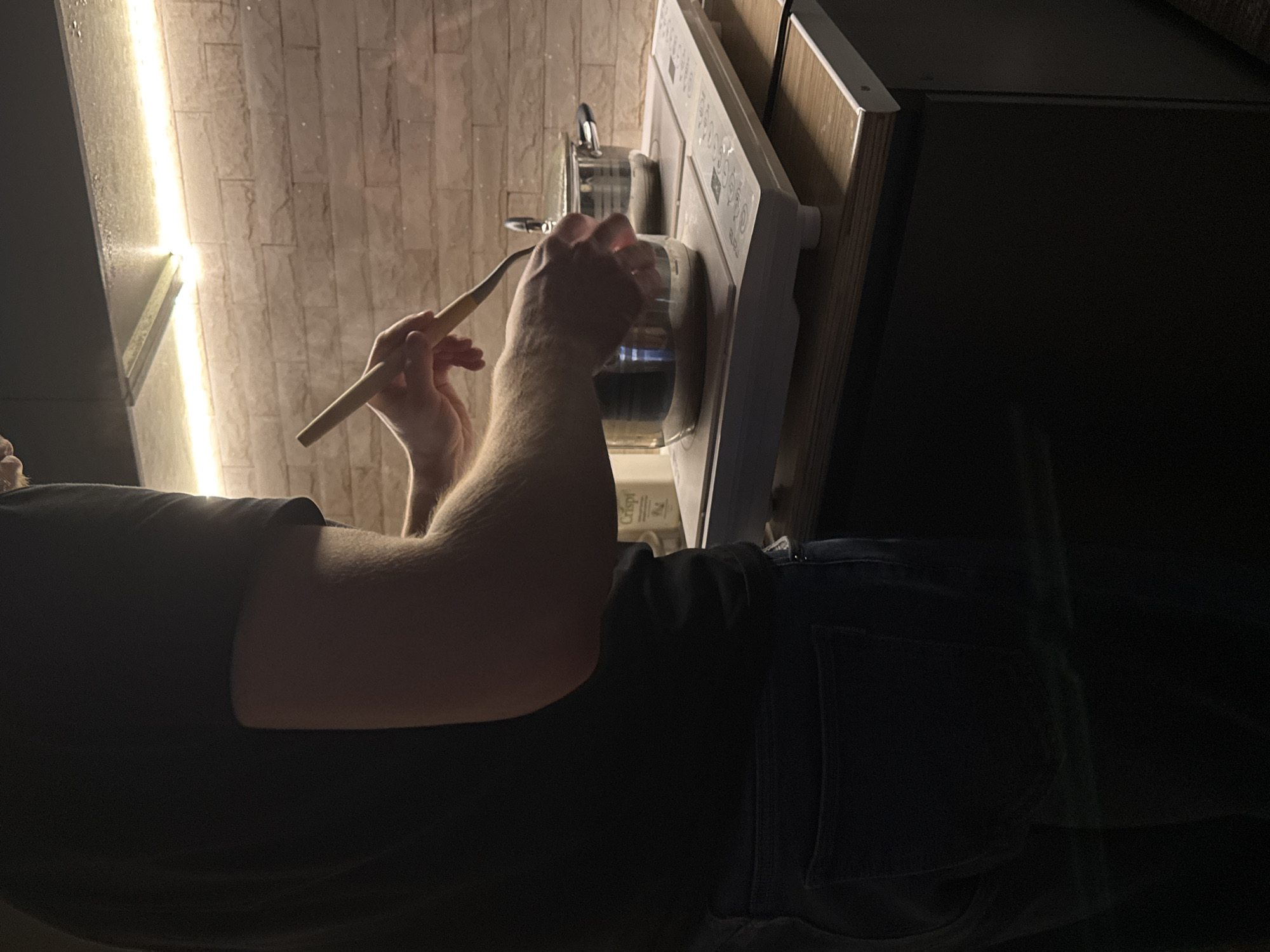 The image depicts a domestic kitchen scene in Perm, Russia, illuminated by warm accent lighting. A male individual, largely obscured from the chest down and facing away from the camera, is present. He wears a dark short-sleeved t-shirt and dark blue jeans. His forearms are visible, and he is actively stirring the contents of a metal pot using a light-colored wooden or bamboo stirring utensil held in his right hand.
The pot rests on a white portable induction cooktop, which features two cooking zones and digital controls. A second, lidded metal pot is also on the cooktop to the right. The cooktop itself is positioned on a light-colored wooden countertop, which appears to be part of a darker, possibly black, lower cabinet unit. The background features a light-colored, brick-patterned wall, above which a strip of warm LED lighting is installed, providing illumination to the cooking area. Above this lighting, a dark, reflective surface is visible, possibly the underside of an overhead cabinet or range hood. A white container labeled "Crispl" is partially visible behind the cooking area on the counter. The overall lighting in the room is dim, with the accent lighting highlighting the immediate cooking area.