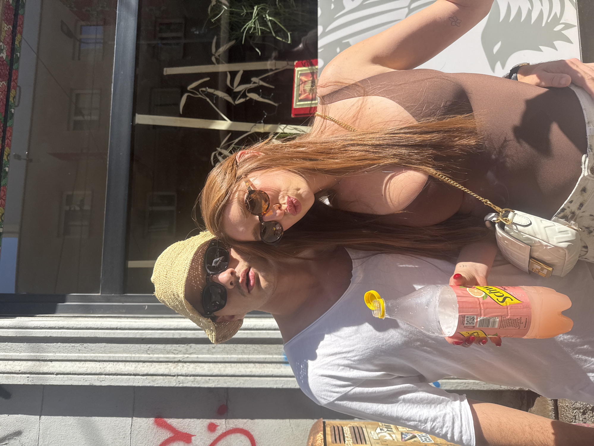 A street-level, medium shot captures two individuals posing for a photo, likely in Milan, Italy. A woman with auburn hair, wearing sunglasses and a brown top with a gold chain, leans her head towards a man sporting a straw hat and sunglasses. He is dressed in a white shirt and holds an open bottle of what appears to be Schweppes soda. The backdrop includes a building facade with a glossy window reflecting the city. Part of a graffiti-covered wall and sidewalk are visible in the foreground.