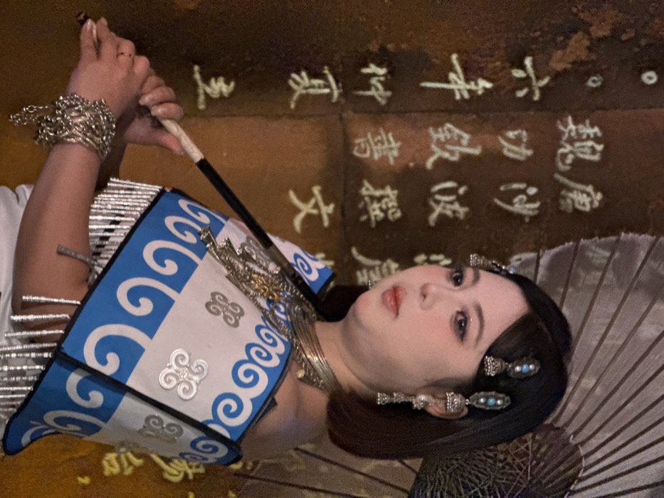 The image shows a woman adorned in elaborate traditional Chinese attire, captured in what appears to be a theatrical or cultural setting, potentially in Yuzhong District, China. The image is rotated 90 degrees clockwise.
The subject is a woman with intricate makeup, including defined eyes and soft red lips, gazing calmly upwards (when the image is viewed upright). Her dark hair is styled back and embellished with ornamental silver hairpins featuring blue and pearl accents. She wears a striking silver stacked bracelet on her left wrist and a multi-layered silver necklace. Her attire includes a blue and white patterned garment, possibly a vest or shoulder piece, adorned with silver spiral motifs. She holds a long, slender, dark-colored prop, possibly a pipe or a handle of another instrument, with both hands. Another prop, resembling an open umbrella, is partially visible to her right.
The background is dark, with classical Chinese characters illuminated or projected onto it, arranged vertically. These characters, when viewed upright, include "魏唐" (Wei and Tang dynasties), "中書撰" (Imperial secretariat compiled/authored), "六年仲夏主" (Sixth year, mid-summer, master/lord), "功飲書" (Meritorious deeds, drinking, writing/books), and "文" (Culture/writing). These texts suggest a historical or literary theme, complementing the traditional costume. The overall impression is one of cultural performance or a staged portrait emphasizing historical or ethnic aesthetics.