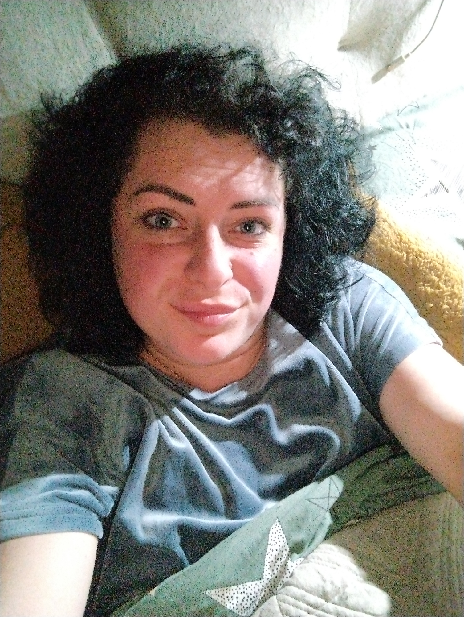 A medium-shot, selfie-style image depicts a woman with fair skin, dark curly hair, and light-colored eyes. She is wearing a gray, draped top. She appears to be lying down on a surface covered with various fabrics and textures, including what looks like a patchwork quilt in shades of green, beige, and white. A yellow pillow or cushion is visible in the upper right corner of the image. The scene is softly lit. There is no evidence or indication that this image was taken in or near Grishanki, Russia.