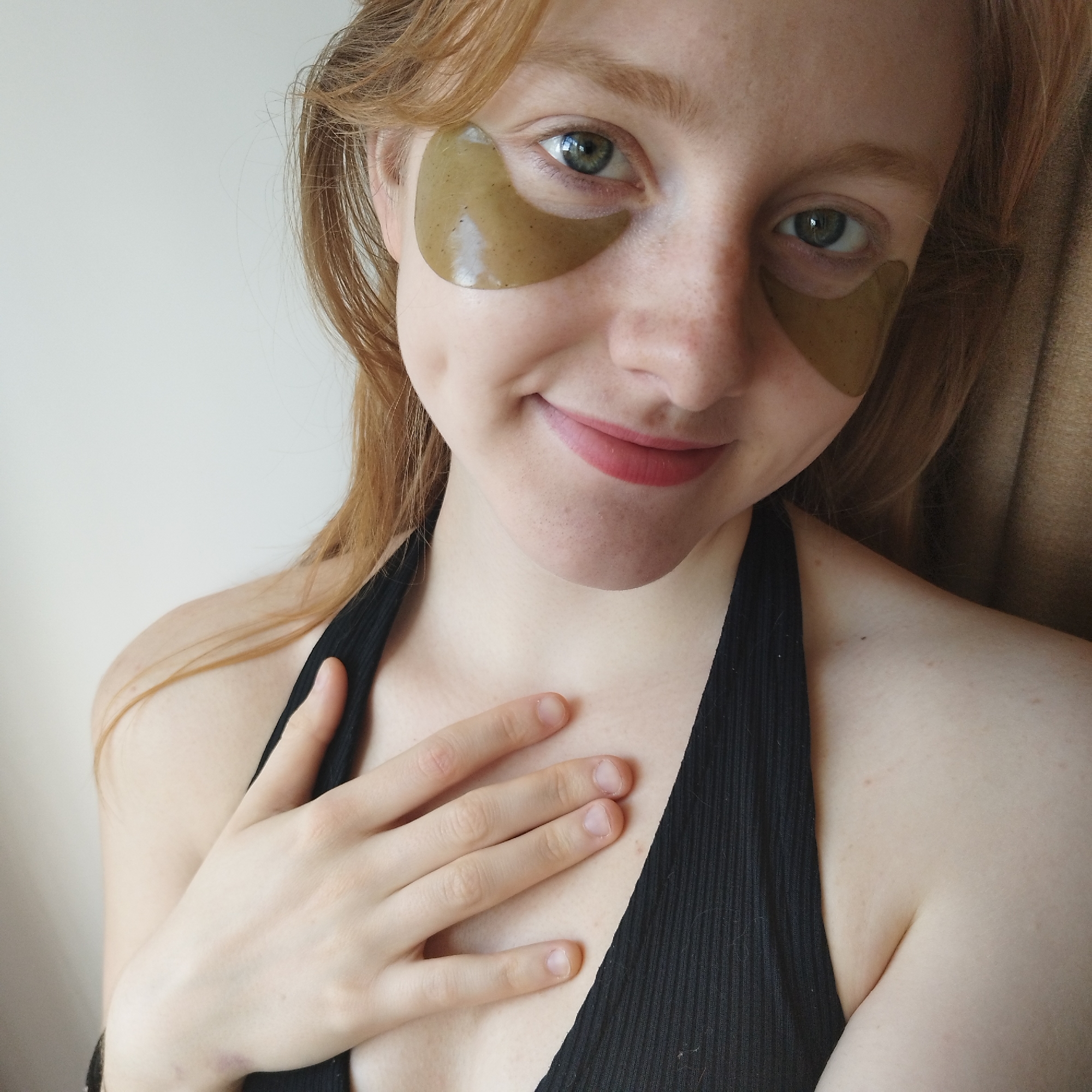 A close-up, brightly lit portrait features a young woman with reddish-brown hair and fair skin, smiling gently at the camera. She has distinct green eyes and a scattering of freckles across her nose and cheeks. Underneath each eye, she wears a translucent, greenish-brown eye mask or patch, suggesting a skincare routine. Her lips are tinted with a light red or pink color, and she exhibits a dimple on her left cheek.
She is wearing a black, ribbed, halter-neck style top or bra. Her left hand is placed on her chest, fingers splayed slightly, and her fingernails appear natural and unpolished. Her hair falls loosely around her face and shoulders. In the background, a plain light-colored wall is visible on the left, and a textured brown or beige fabric, possibly a curtain or blanket, can be seen on the right side of the frame. The lighting suggests an indoor setting, likely during the daytime. The overall mood is relaxed and pleasant. There is no visible text in the image.