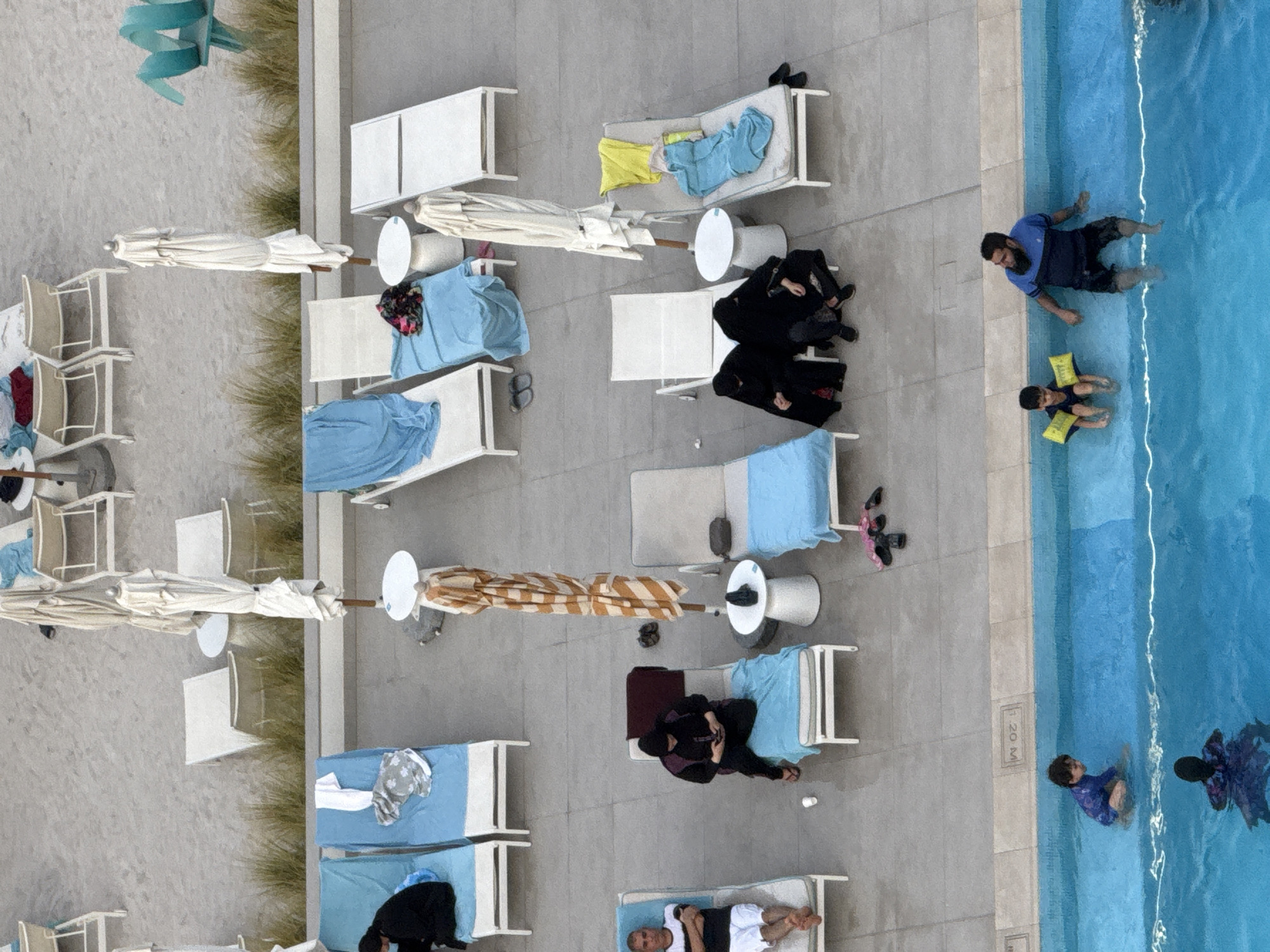 This is an aerial, high-angle, outdoor, full shot photograph taken at the Waldorf Astoria Ras Al Khaimah in the United Arab Emirates. The scene captures a resort area with a pool and a sandy beach in the background. Several lounge chairs, some occupied, are arranged around the pool, with umbrellas providing shade.
In the pool, a young child in a blue swimsuit and yellow arm floats is playing. To the right, an adult male in a blue t-shirt and dark shorts, also partially in the water, is seated at the edge of the pool. Another child, wearing a purple and black swimsuit, is in the deeper part of the pool, visible from the shoulders up.
On the lounge chairs near the pool, a man in a black and white striped t-shirt and white shorts reclines on a blue-covered chair. Another person in a dark burqa and headscarf is seated on a blue-covered chair, looking at their phone. Further to the right, two women, both wearing black burqas and headscarves, are seated on a white lounge chair. A collection of beach towels and umbrellas are scattered around the lounge chairs.
In the background, beyond a strip of greenery, the sandy beach area is visible with more lounge chairs and umbrellas. Some of the beach chairs are occupied by people in modest attire, suggesting a family-oriented resort. The overall impression is a relaxed resort setting, likely during daylight hours.