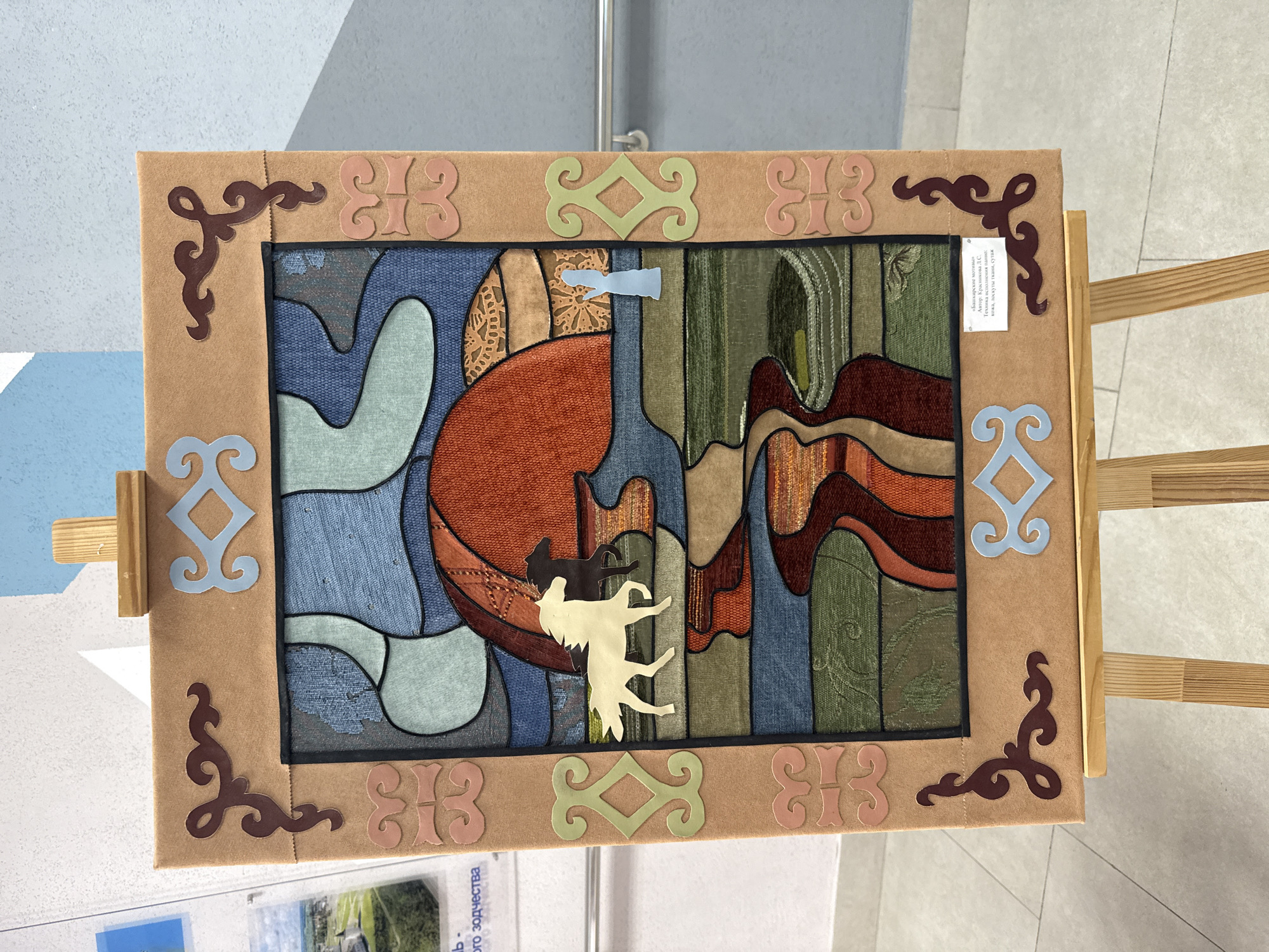 The image shows a framed piece of art displayed on a wooden easel. The art is made of various fabrics and materials sewn together to create an abstract scene with animals. The frame is decorated with stylized, traditional-looking ornamentation in different colors. A small information card is attached to the right side of the frame, with text in Russian indicating the name of the piece "Bashkir motives", the author L.S. Krasnikova, and the materials and technique used, which includes skin, scraps of fabric, and braid. The easel is positioned in front of a wall, with a hint of a poster or brochure visible in the foreground. The location is implied to be in Tobolsk, Russia, as indicated by the text on the card, which mentions the "heritage of architectural design" possibly referring to a cultural organization or location in Tobolsk.