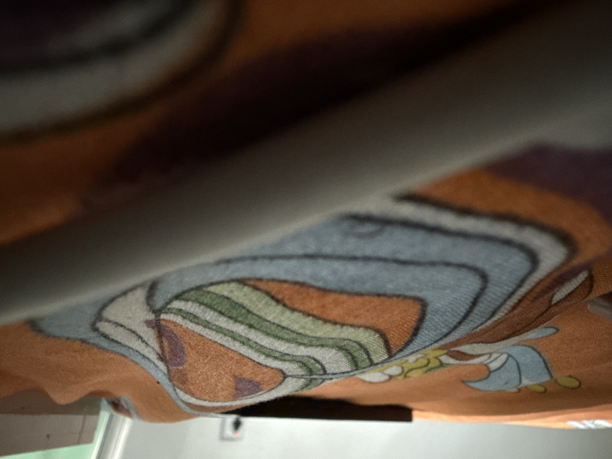 The image shows a close-up, low-angle view of a patterned fabric, possibly a blanket or tapestry, with an abstract design featuring concentric wavy lines in shades of orange, blue, green, and purple. A dark, rectangular object, resembling a book or electronic device, is placed horizontally on the fabric in the upper background. The fabric is draped over a surface, creating folds and shadows. The overall lighting is dim, with a brighter light source from the left, casting shadows on the fabric. The background is out of focus, showing a plain wall and a hint of a window with a green tint. The scene appears to be indoors, but no specific location in Uyo, Nigeria, can be determined from the visual information provided. There are no people, actions, or interactions visible in the image.