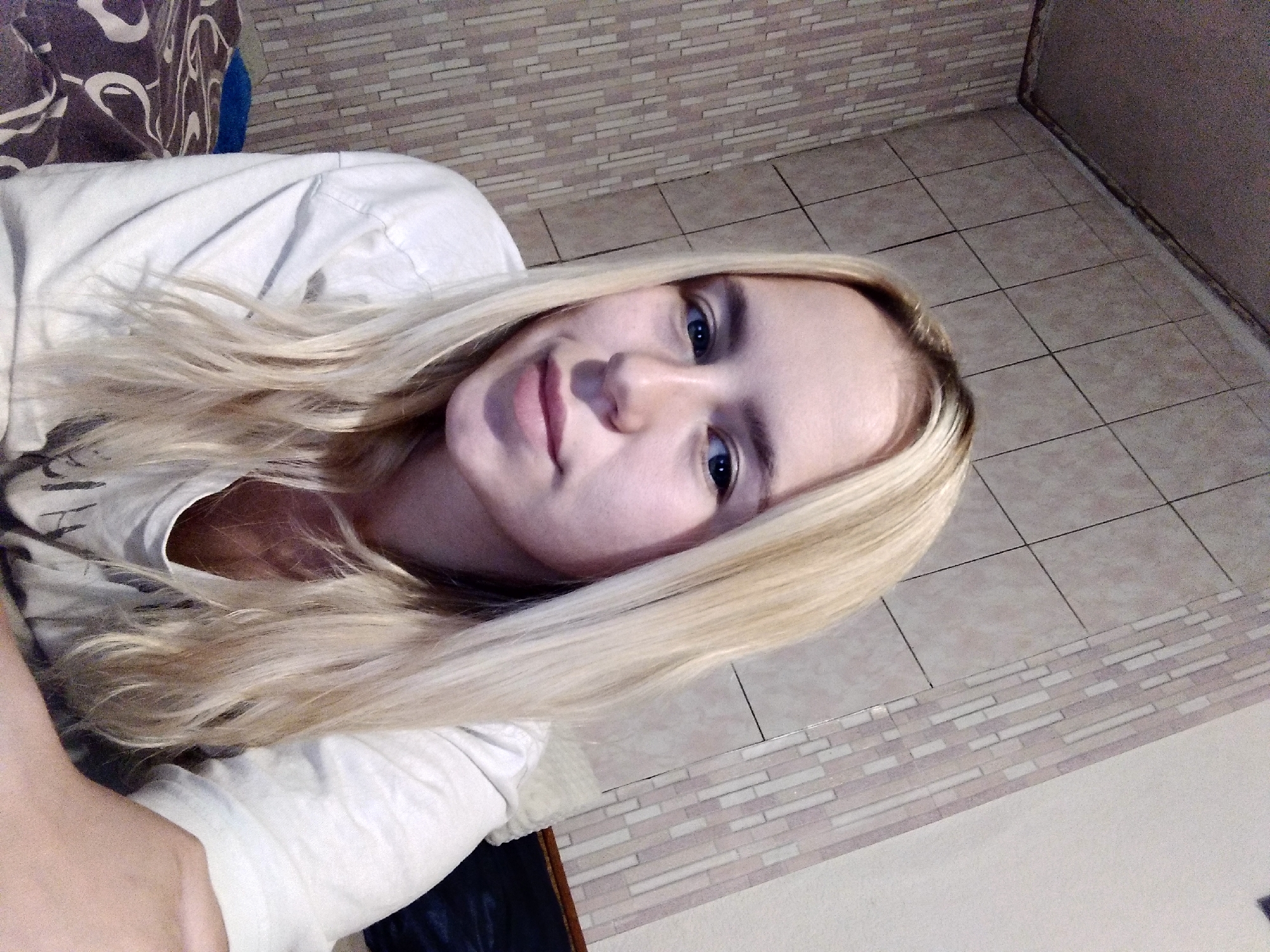 A young woman with blonde hair and blue eyes is pictured in a close-up, head-and-shoulders shot. She is wearing a white t-shirt and looking to the side. The background consists of tiled walls and a bed. The lighting appears to be artificial and dim. The image was taken indoors, likely in a bedroom, and the location is unknown.
