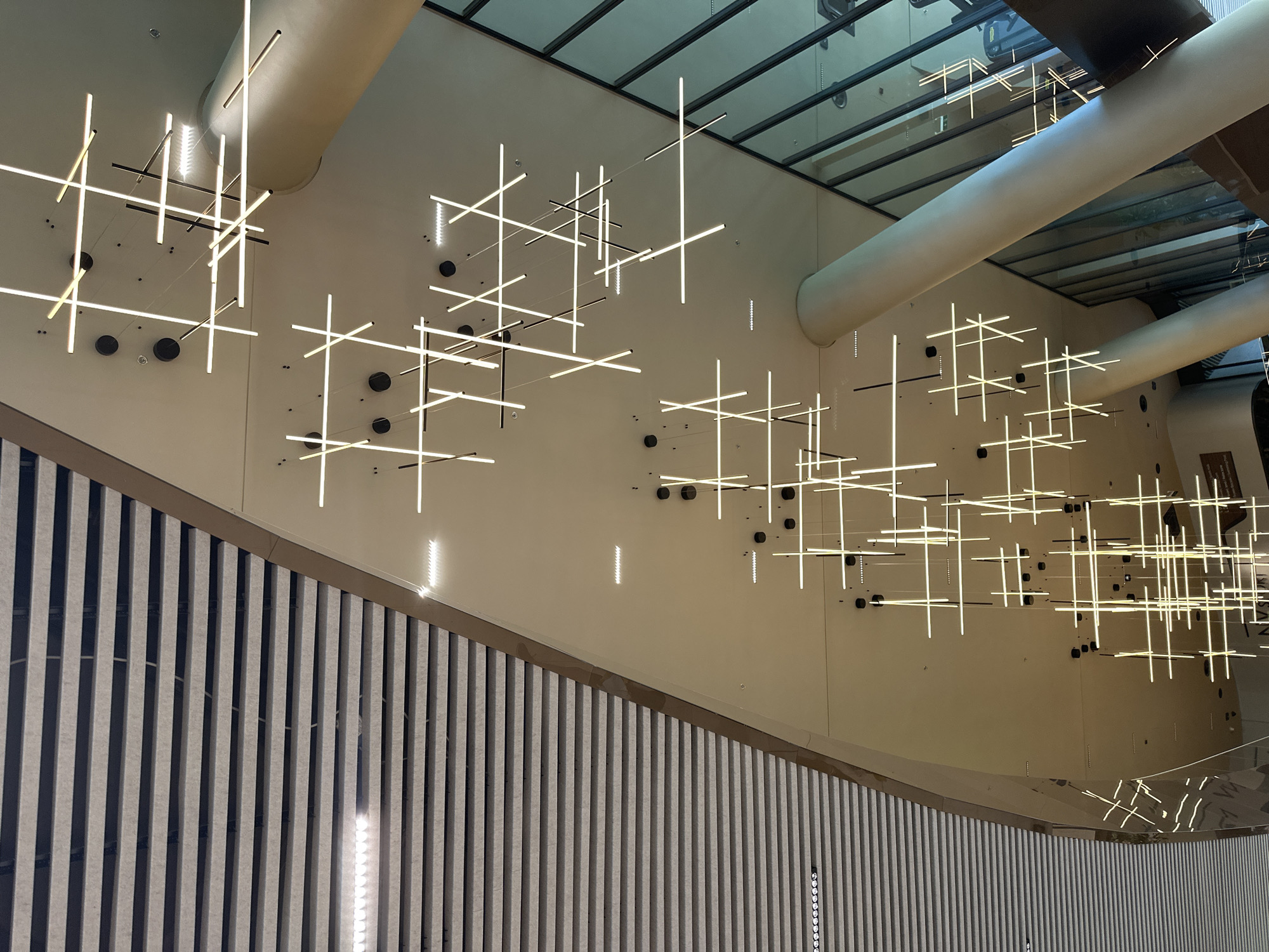 This media file captures an interior scene featuring a modern architectural design with striking lighting installations. The primary subjects are a series of linear, white LED lights suspended from the ceiling, arranged in abstract, intersecting patterns that resemble constellations or geometric sculptures. These lights illuminate a light-colored, slightly curved wall that dominates the background.
Below the illuminated wall, a textured surface with vertical gray and white stripes extends upwards, creating a visual contrast with the smooth, painted wall above. This striped surface appears to be a decorative element, possibly a wall paneling or a balustrade.
The ceiling is partially visible, featuring a reflective glass panel with dark framing, suggesting a modern atrium or a large indoor space with natural light filtering through. Two large, smooth, cylindrical structures, possibly air ducts or structural elements, are also present in the upper portion of the image, adding to the architectural complexity.
The lighting suggests that the scene is likely indoors and illuminated, possibly during daylight hours given the ambient light and reflections. There are no people visible in the media file. The overall impression is one of contemporary design, artistic lighting, and spaciousness. The location context provided is Sofia, Bulgaria, which implies the setting is within a building in that city. No visible text is present.