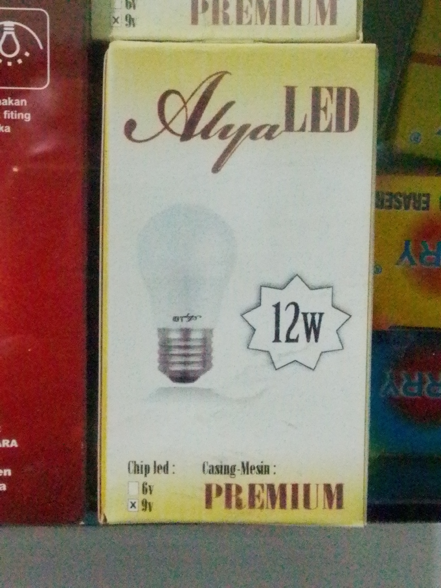 The image is a close-up, eye-level shot of packaging for an LED light bulb, likely taken in a store or warehouse in Bandar Lampung, Indonesia. The packaging is primarily yellow and white, with brown text and graphics. Prominently displayed is the brand name "Alya LED" in a cursive, stylized font. Below the brand name, a graphic of an LED light bulb is shown. To the right of the bulb graphic, the wattage is indicated as "12W" within a starburst shape. At the bottom of the packaging, there are details about the "Chip led" and "Casing-Mesin," with options for "6v" and "9v" indicated by checkboxes, where "9v" is marked with an 'x'. To the left of the LED packaging, a portion of a red box with white text is visible, with the words "bakan," "fiting," and "ka" discernible. In the background, parts of other product packaging are visible, including a green box with yellow text and a blue box with red and yellow graphics, suggesting a retail environment. The lighting is somewhat harsh, creating a slight glare on the packaging. The overall scene is static, focusing on the product itself.